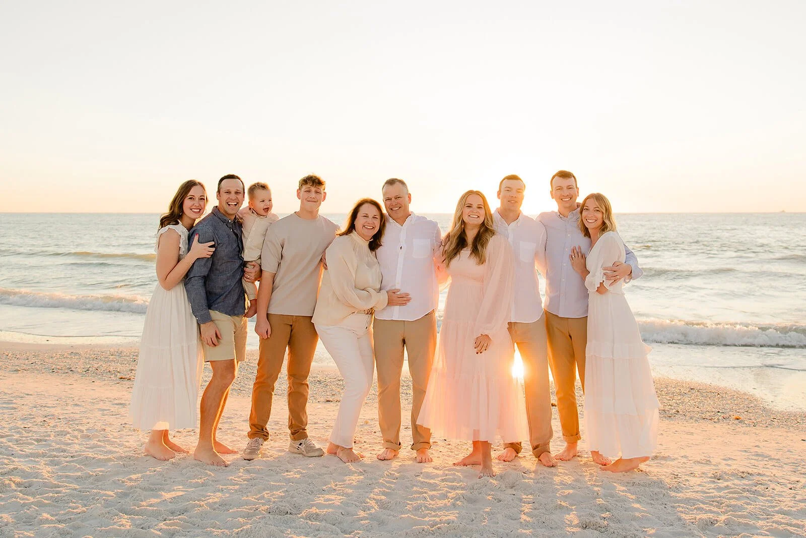 A family of ten standing barefoot on the beach during sunset, dressed in light-colored clothing, smiling and embracing each other photographed on a Naples Florida beach.