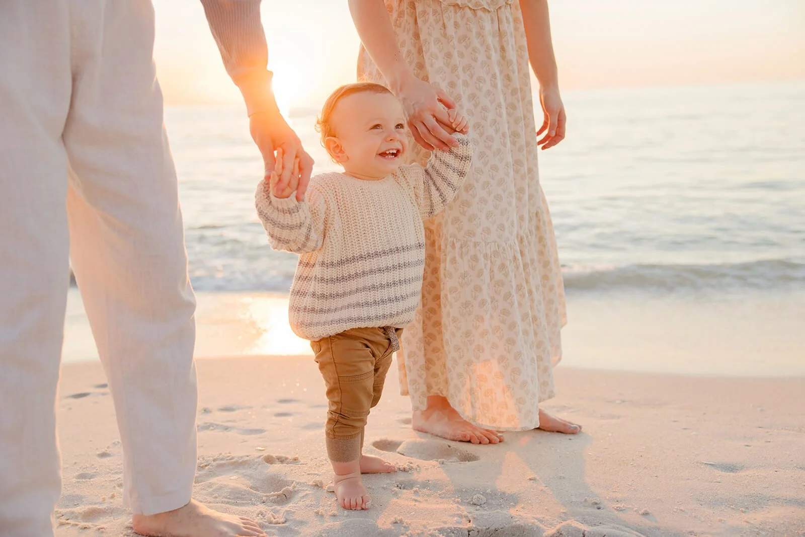 A smiling toddler holding hands with two adults on a beach at sunset, with the ocean in the background during a sunset beach portrait session in Naples FL.