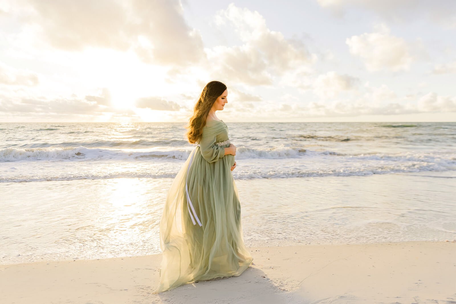A woman in a flowing green dress standing on a beach at sunset, with the ocean and cloudy sky in the background during a maternity session on a Naples, Fl beach.