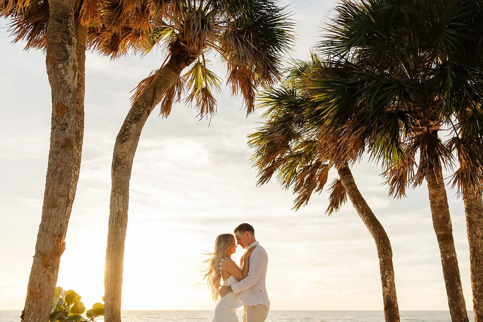 A couple dressed in white embracing on the beach at sunset, with tall palm trees in the foreground and the ocean in the background in a Naples, Fl engagement session.