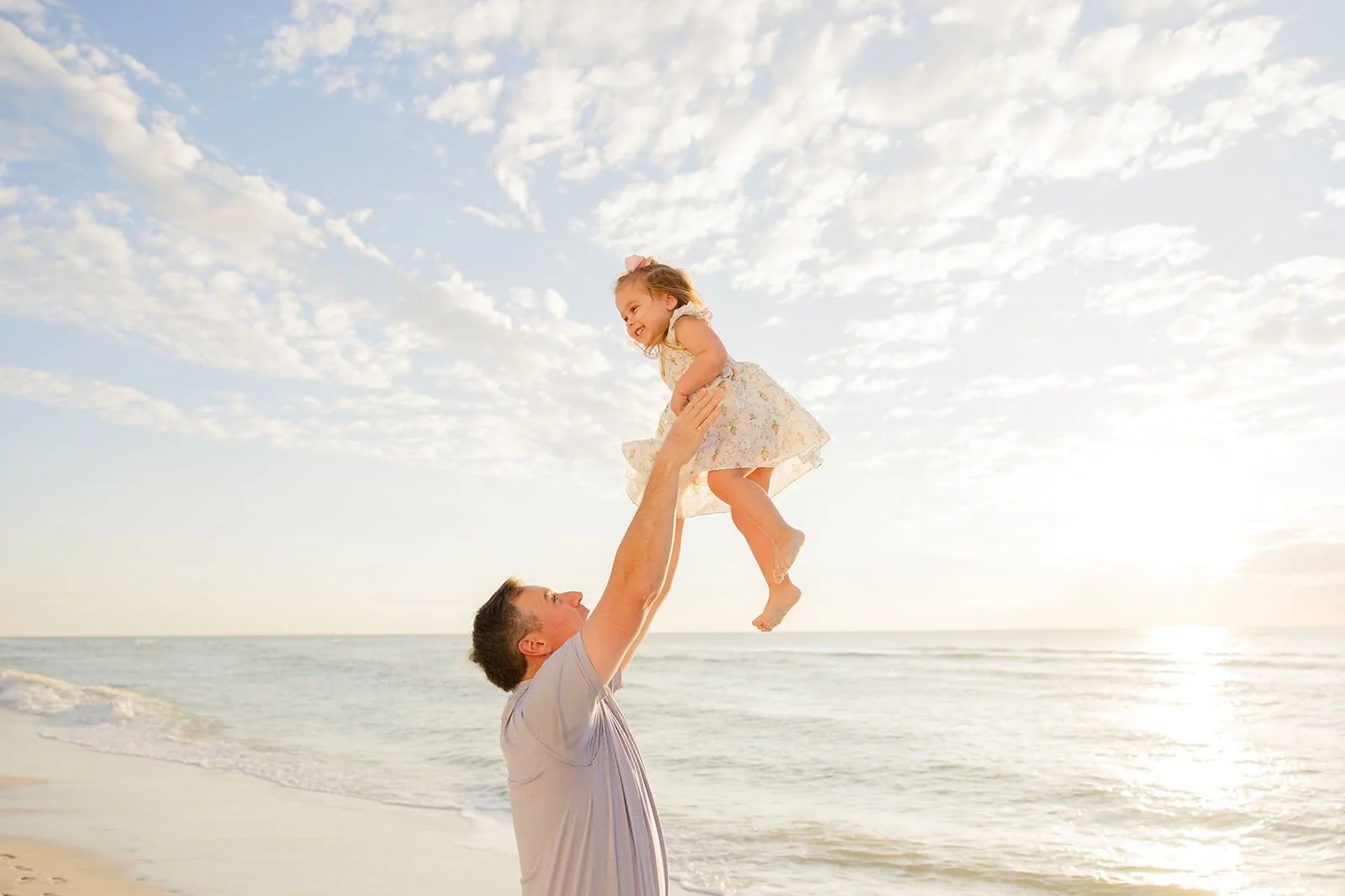 A father lifting a young girl in the air on a beach at sunset, with the ocean and sky in the background during family portraits on Naples Florida beaches.