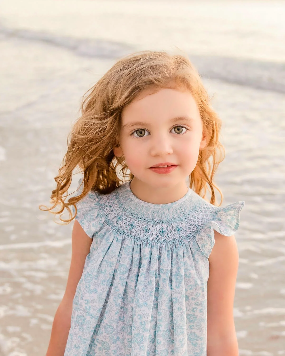 A young girl with red hair and green eyes standing on a beach, wearing a light blue dress with ruffled sleeves, with the ocean in the background.