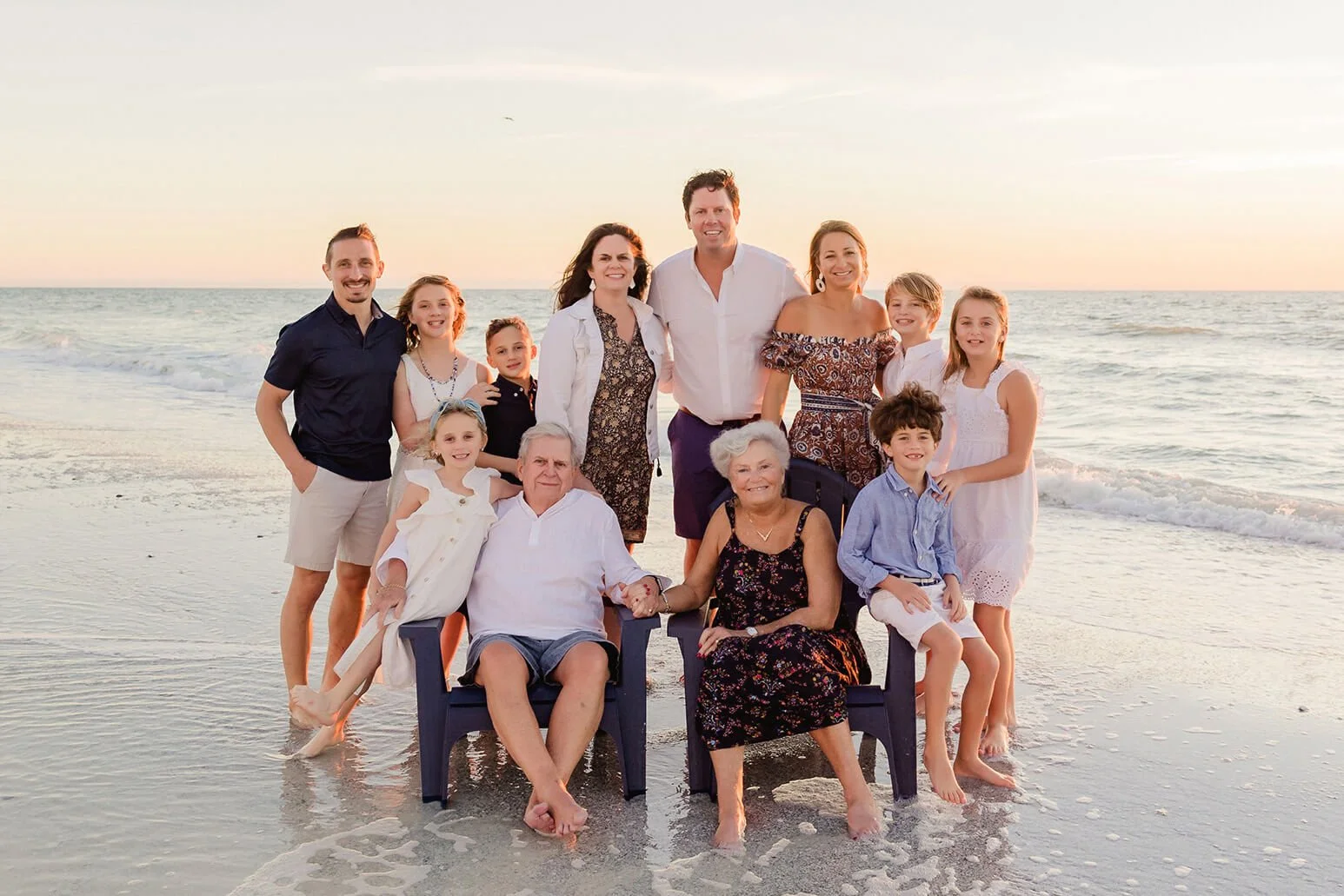Family of fifteen posing on a sandy beach at sunset, some sitting on chairs and others standing, with ocean waves in the background for a family photo in Naples, Florida.