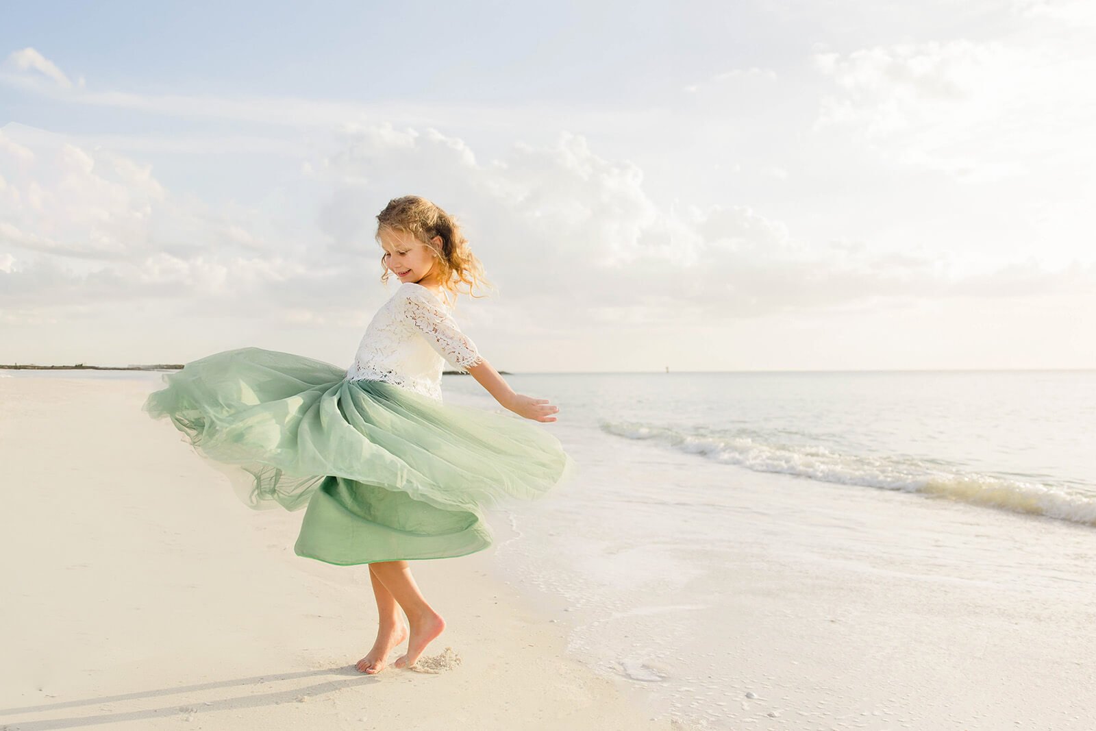 A young girl wearing a light green skirt and a white lace top twirling and playing on a sandy beach near the water with clouds in the sky during a family photo session in Naples Florida.