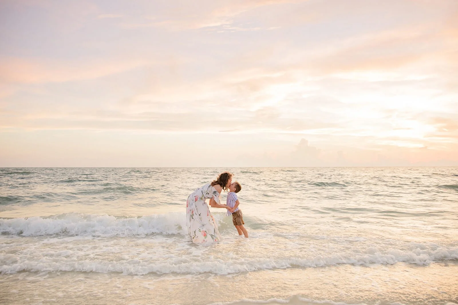 A mother kisses her son standing in the ocean waves at sunset during Naples beach family portrait session.

