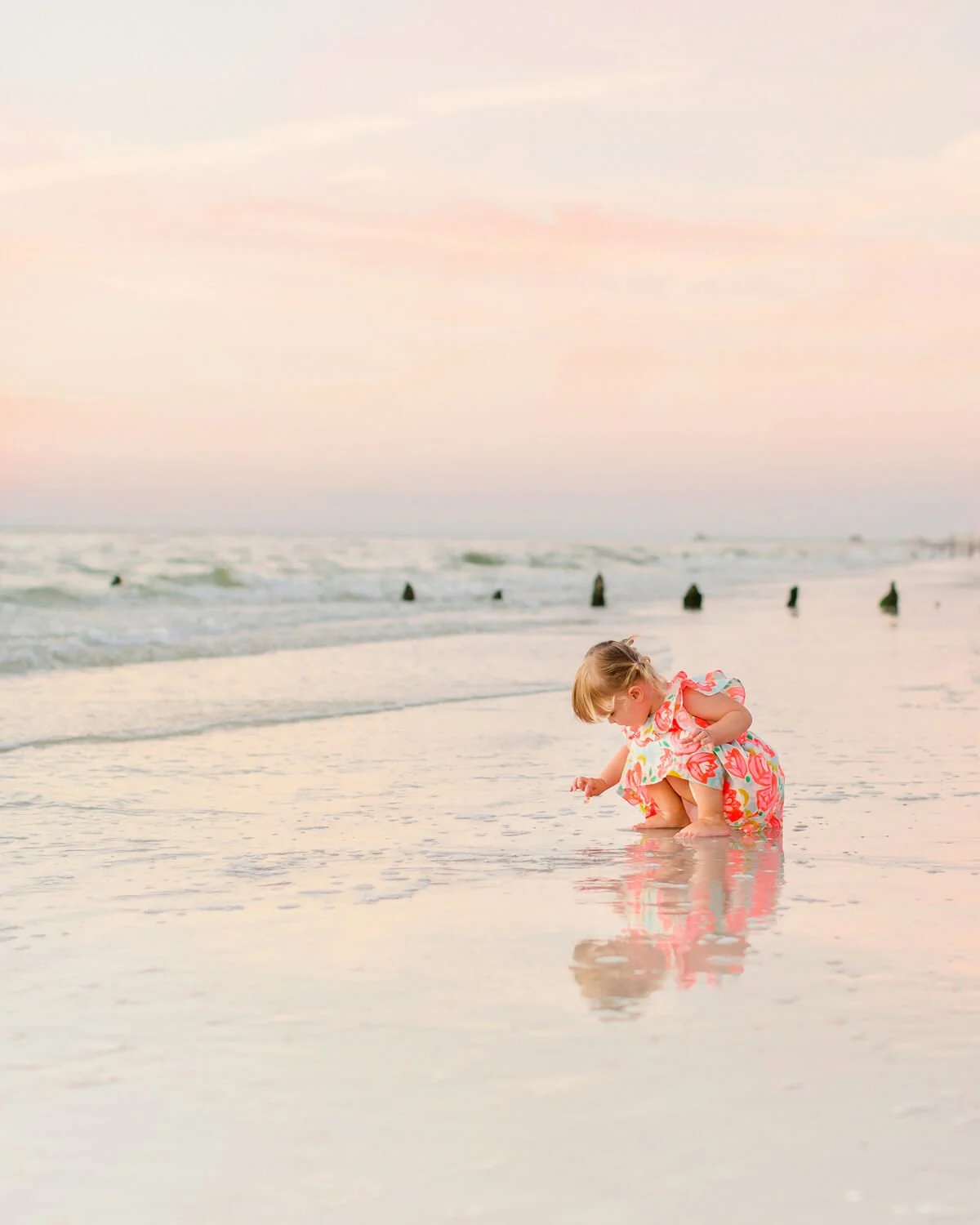A young girl in a colorful floral dress squatting on the wet sand at the beach, playing with the water during sunset, with the ocean and sky in the background during a Naples beach family portrait session.