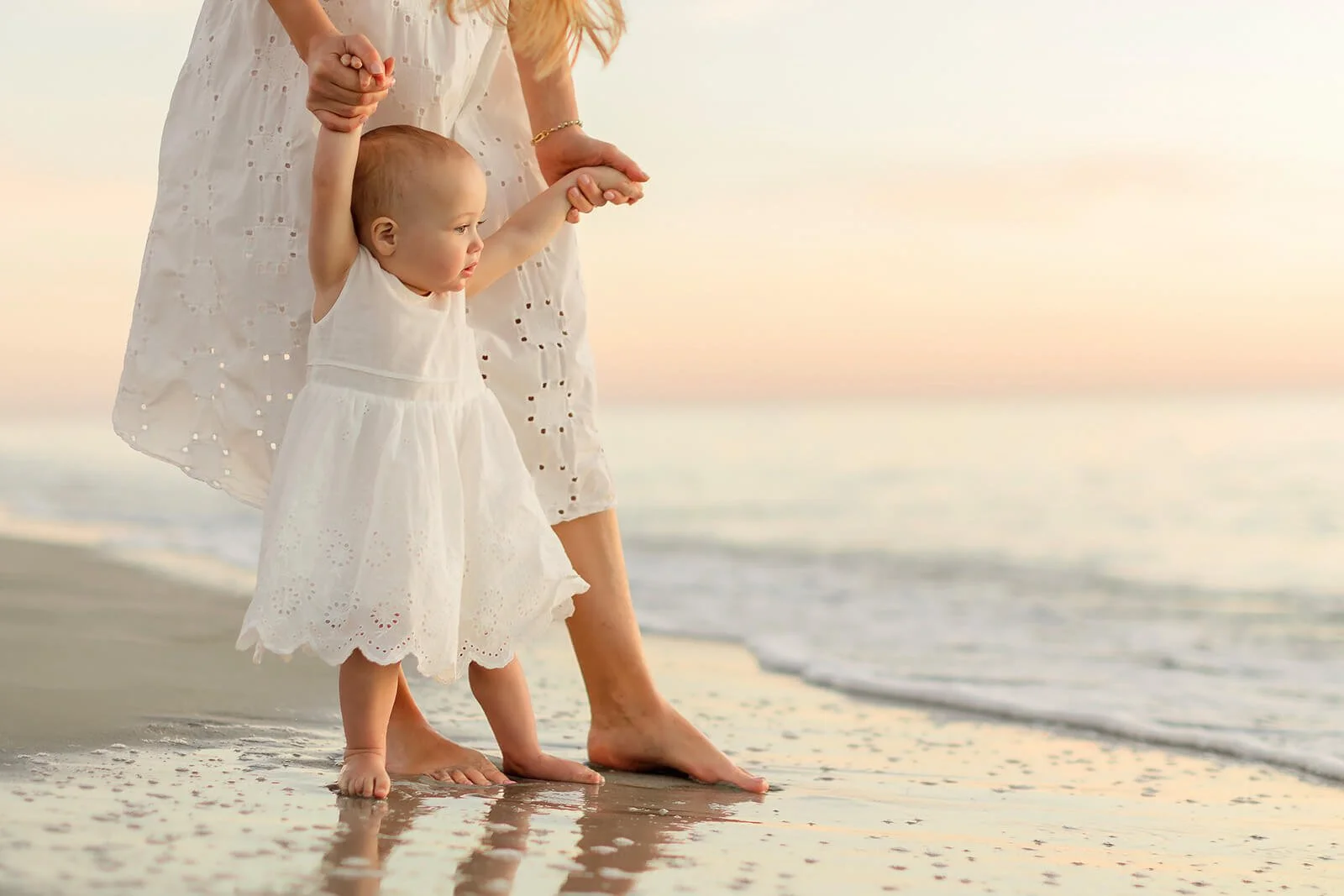 A mother helps her baby daughter walk along the shoreline at sunset, holding the girl's hand as she steps into the water during sunset beach portraits Naples Florida.
