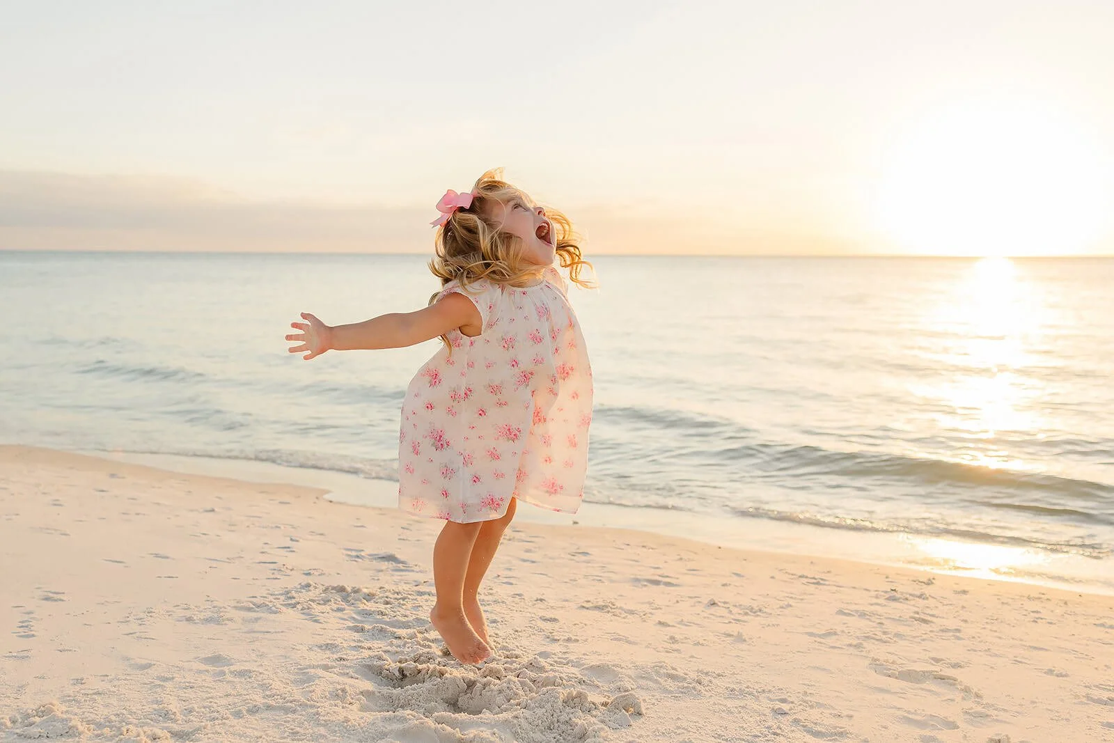 A young girl with blonde hair wearing a light pink dress with pink flowers, standing barefoot on a sandy beach at sunset, arms outstretched, smiling and laughing with her head tilted back while at a sunset beach portrait session in Naples FL.