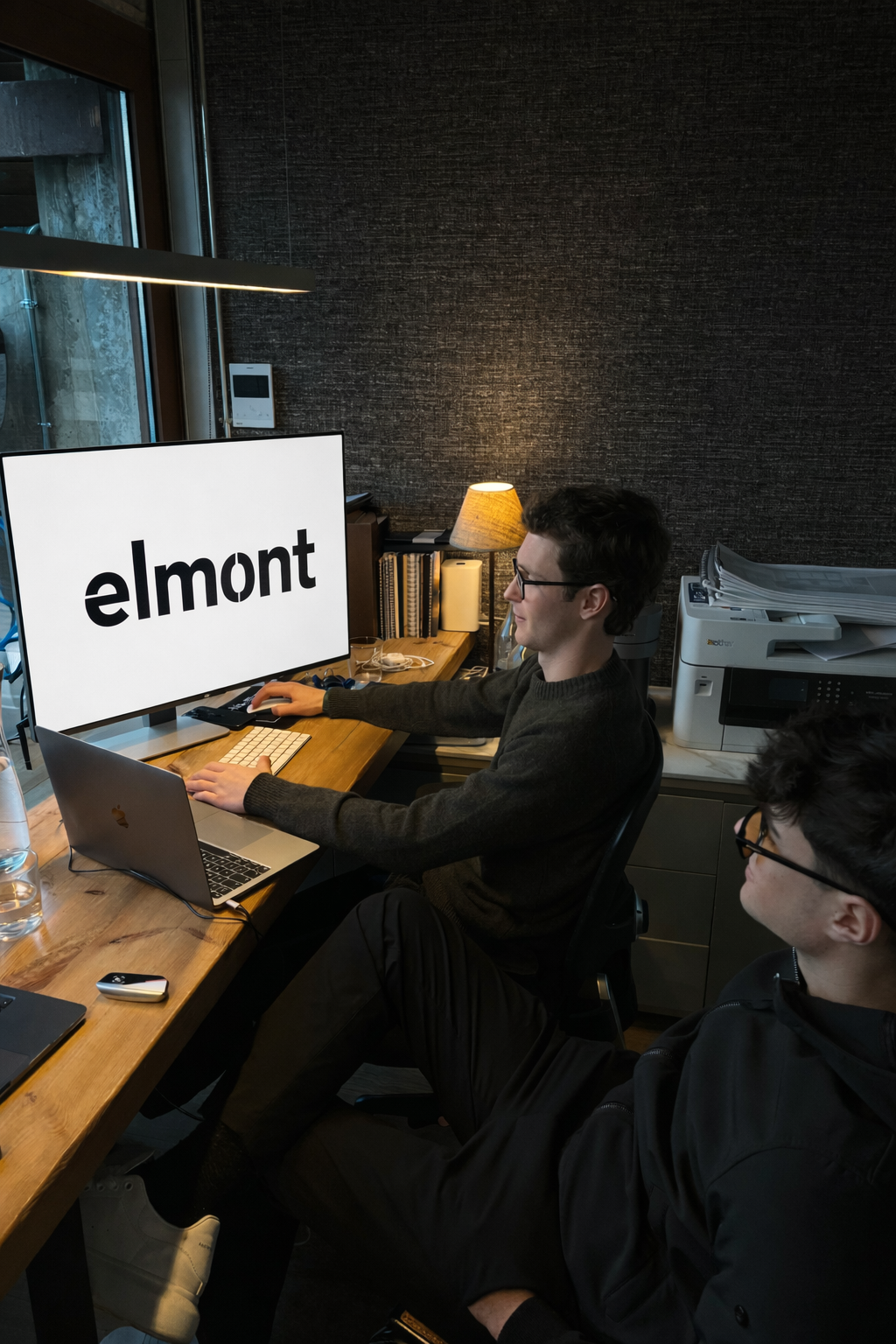 Two young men with glasses sitting at a wooden desk in a modern office, working on a computer with a large screen displaying the word 'elmont'. One is typing on a keyboard, while the other observes. The office has a dark textured wall, a window, a lamp, a printer, and books on a desk behind them.