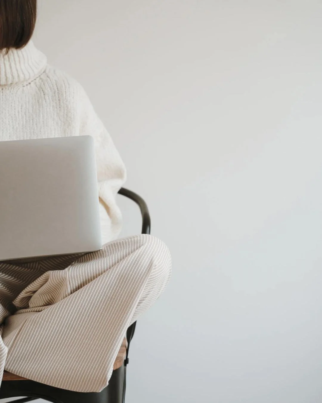 A person sitting on a black chair with a brown seat cushion, wearing a white turtleneck sweater and beige pinstripe pants, holding a silver laptop against a plain white wall background.
