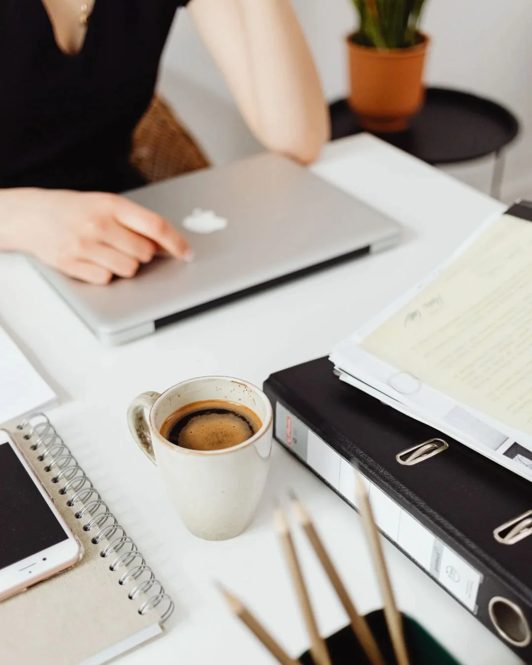 Desk with coffee cup, notebook, smartphone, binder, papers, and a person working on a laptop.