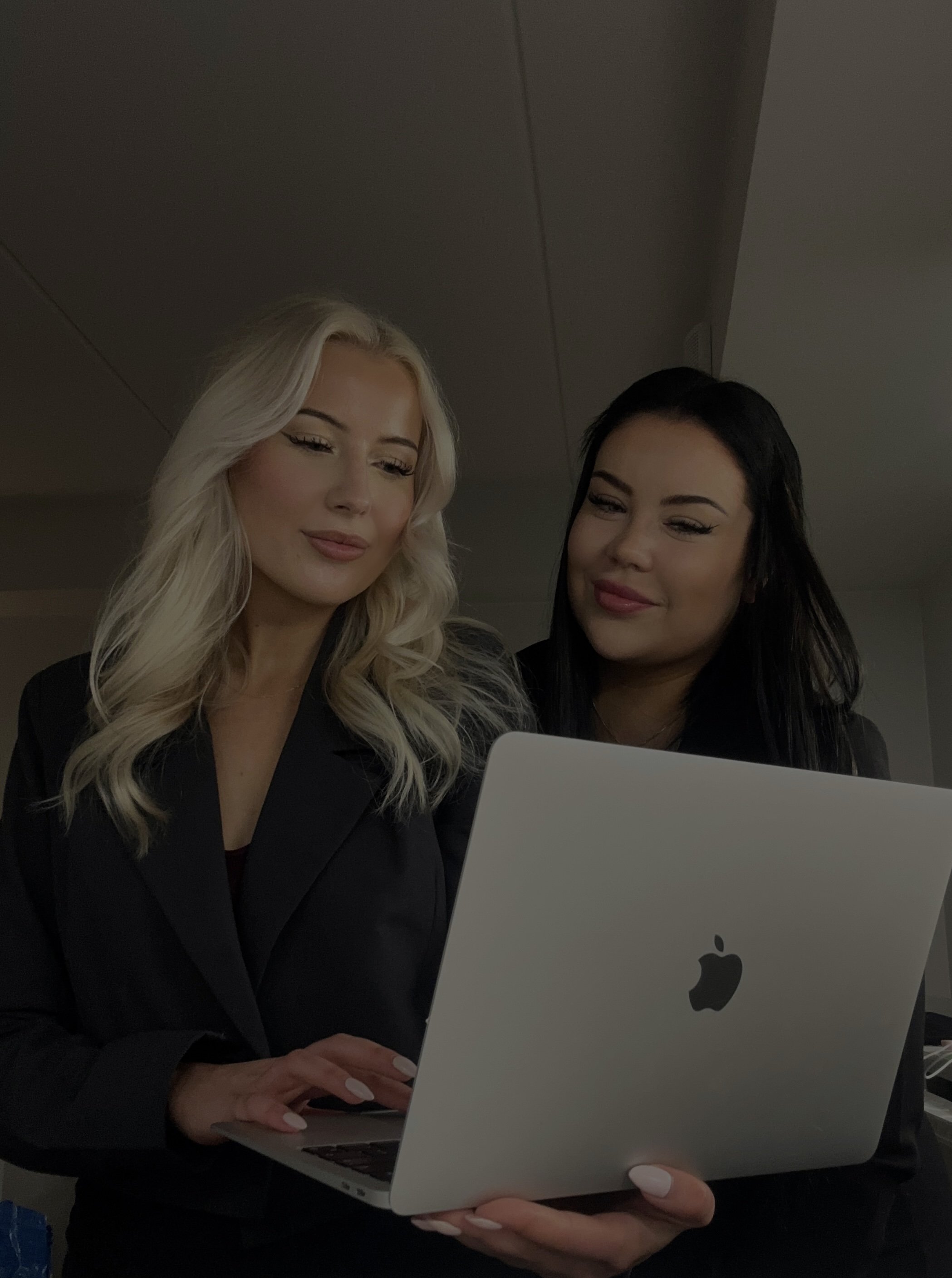 Two women, one with blonde hair and one with black hair, looking at a silver Apple MacBook, standing indoors with a ceiling in the background.