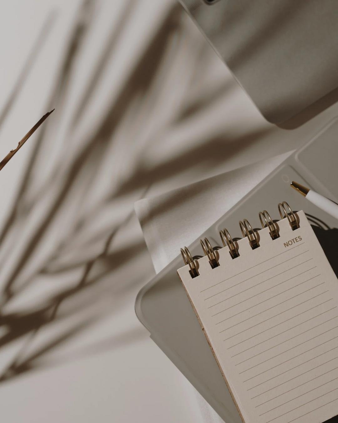 An open lined notepad with 'NOTES' written on the top, placed on a white surface among various papers, a white pen with gold accents, and a gold-tipped paintbrush.