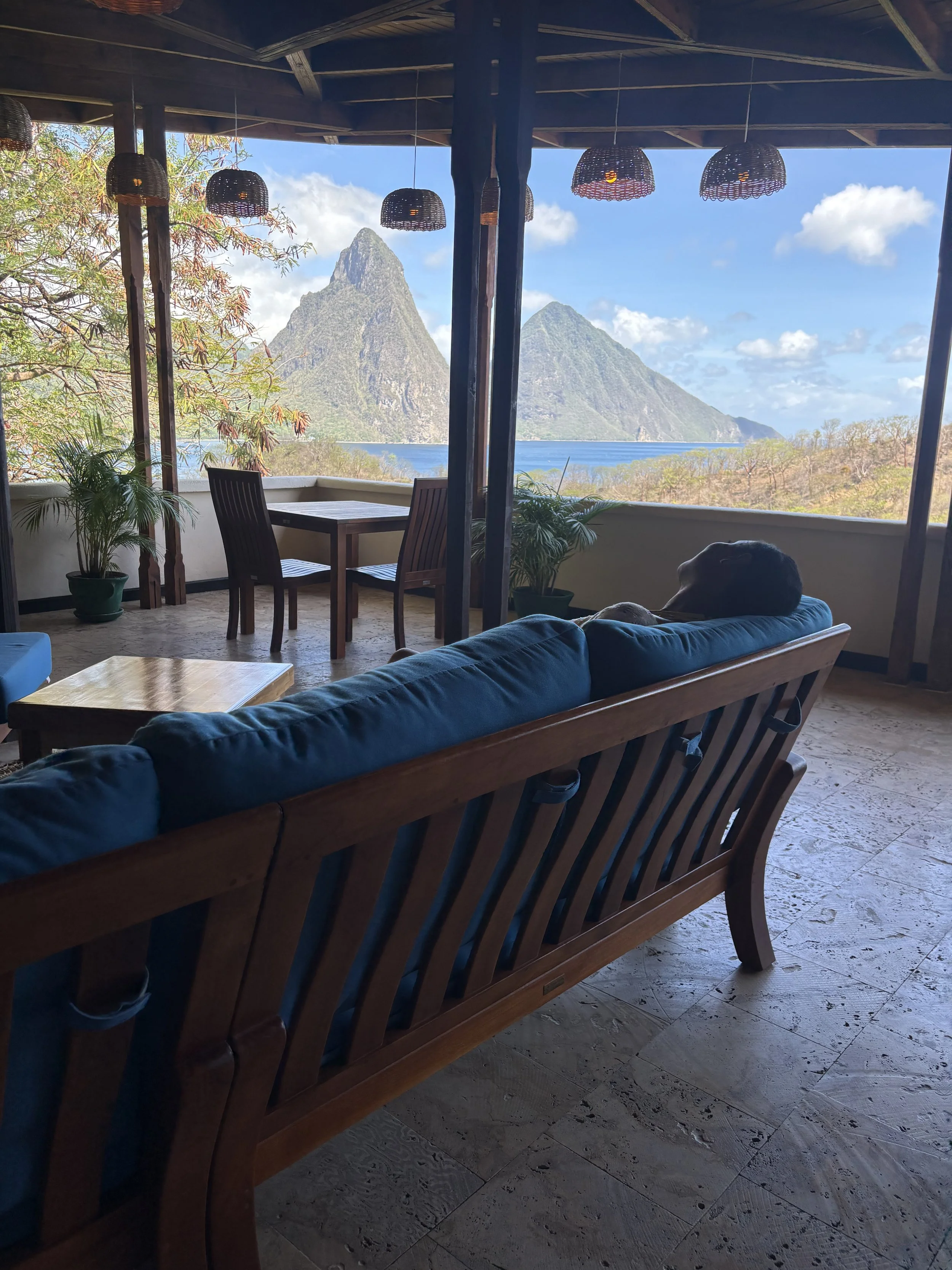 Indoor sitting area with a couch and table, overlooking mountains and a body of water through large open windows, with hanging lamps and potted plants.