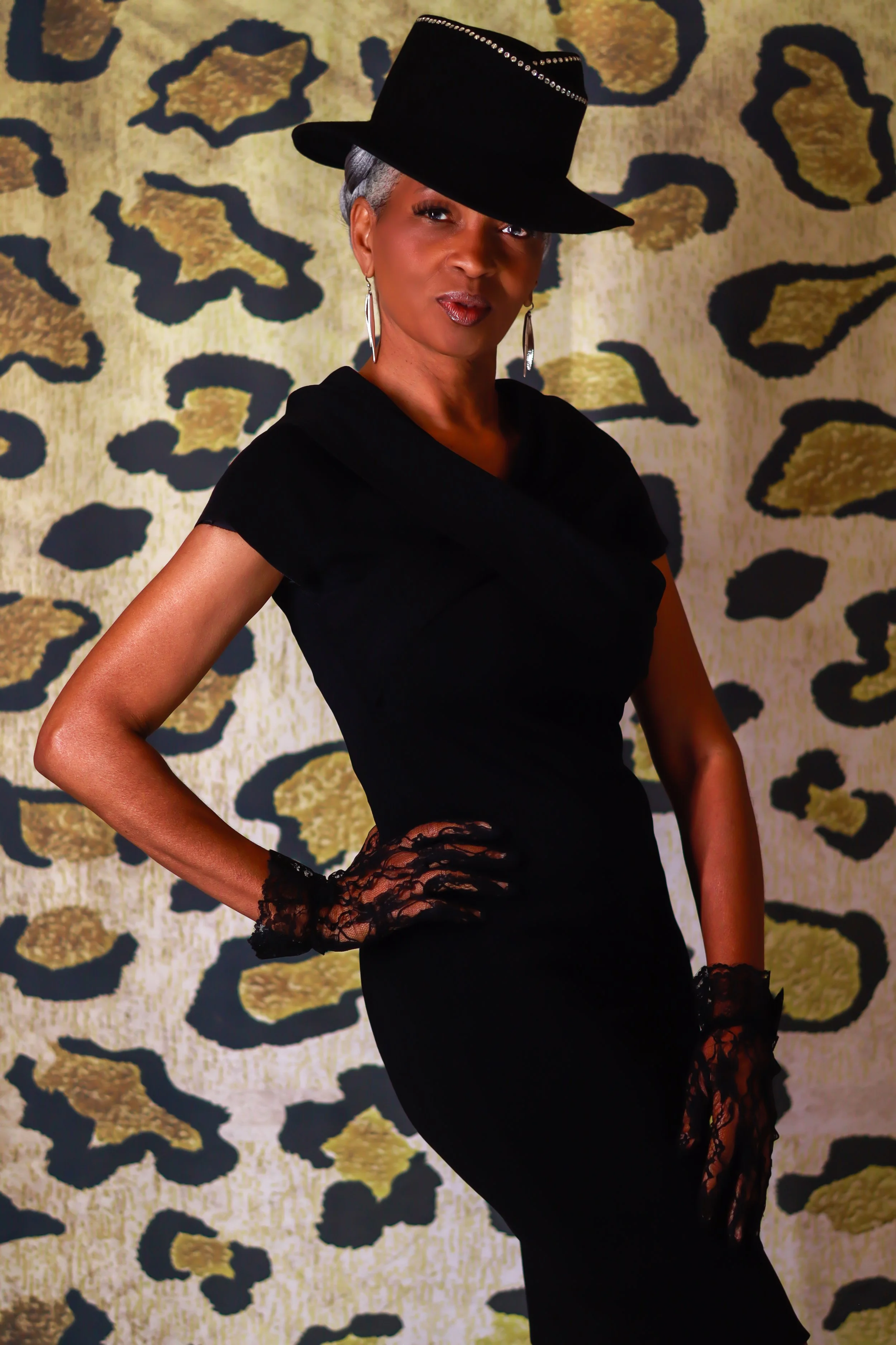 A confident woman in a black dress and lace gloves poses in front of a leopard print background, wearing a stylish black hat with a decorative band.