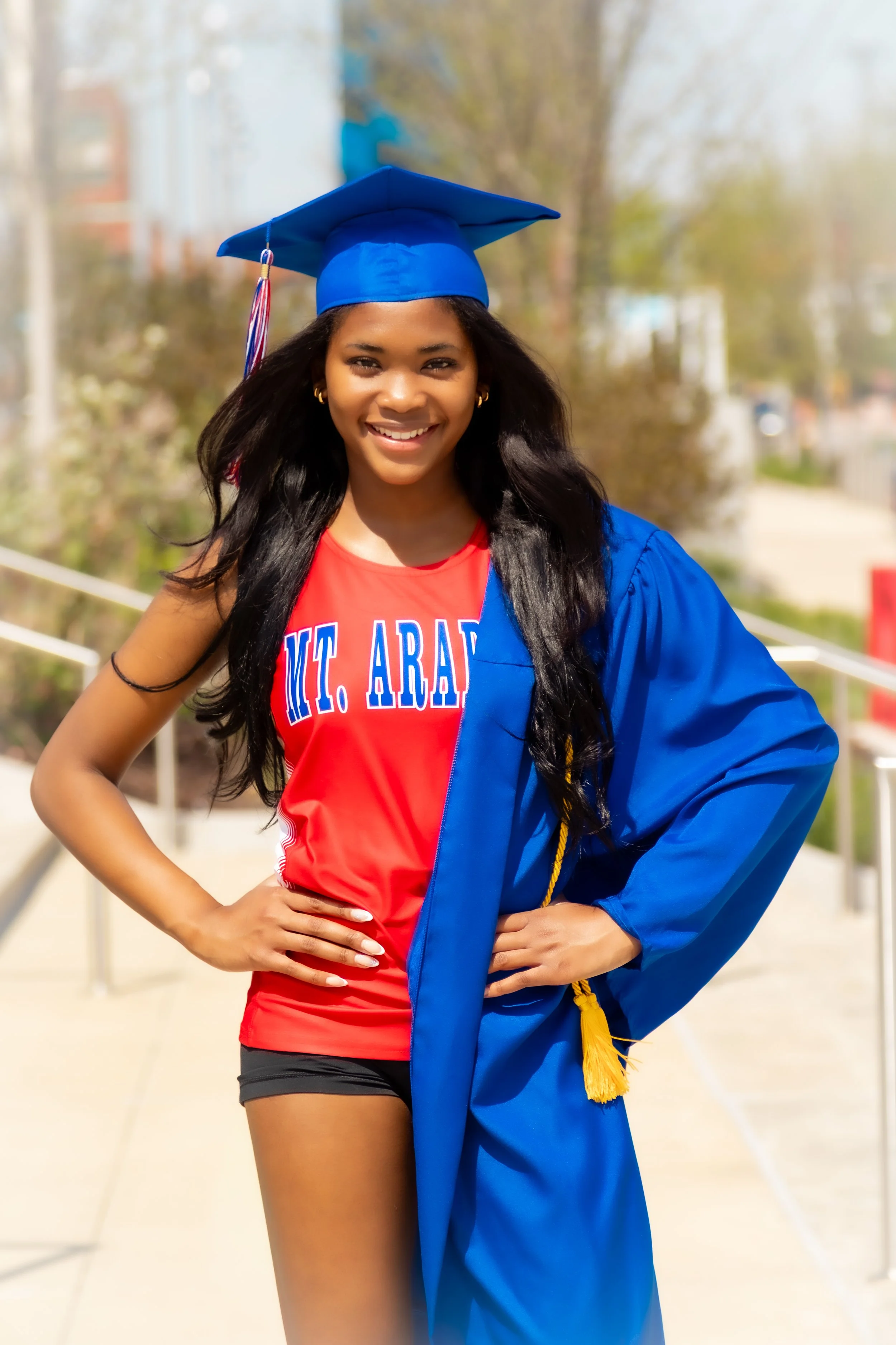 A young woman in a red shirt and black shorts, wearing a blue graduation cap and gown, standing outdoors with a bright smile.