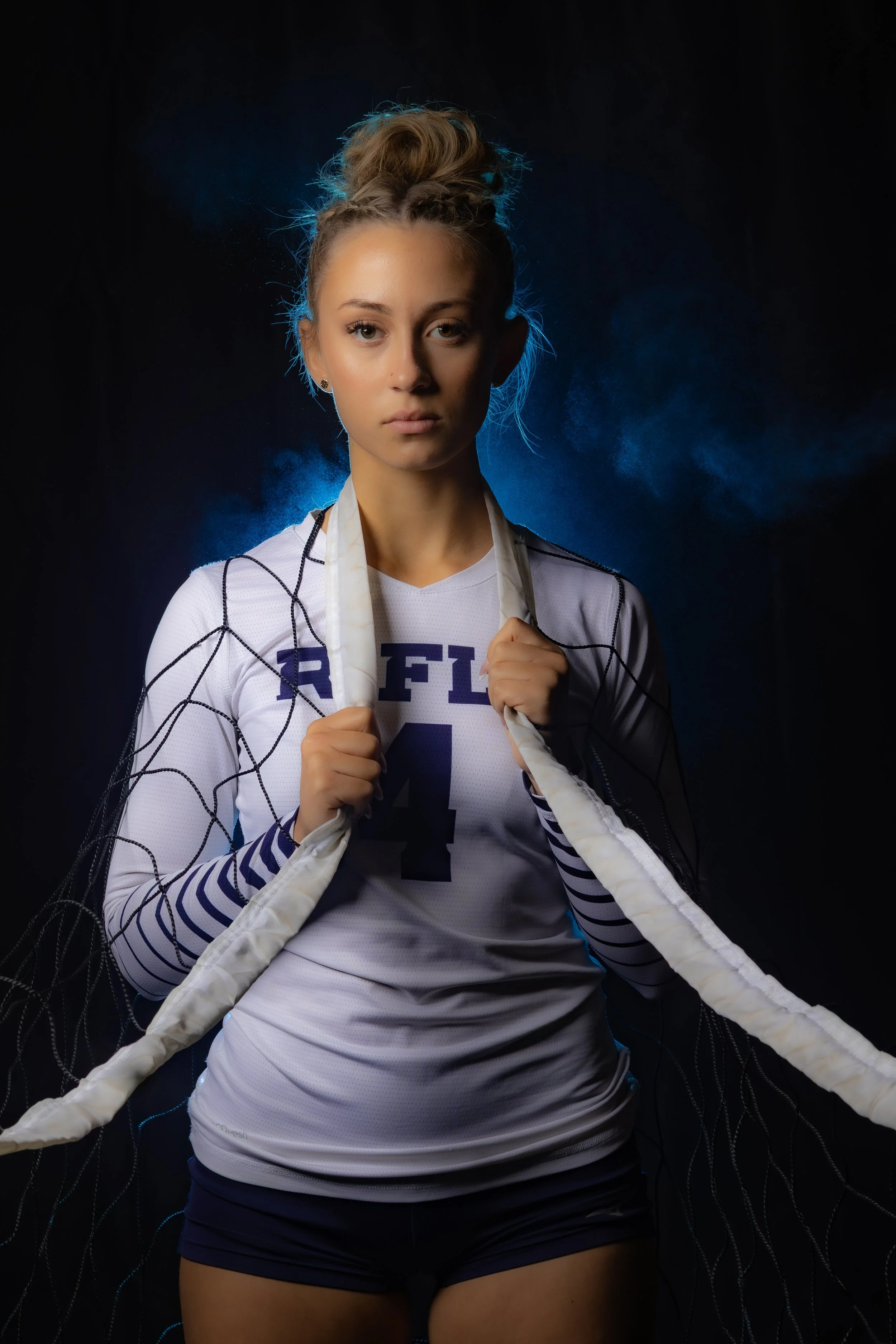 Young female volleyball player holding a net, wearing a white jersey with the number 4, standing against a dark background with blue lighting.