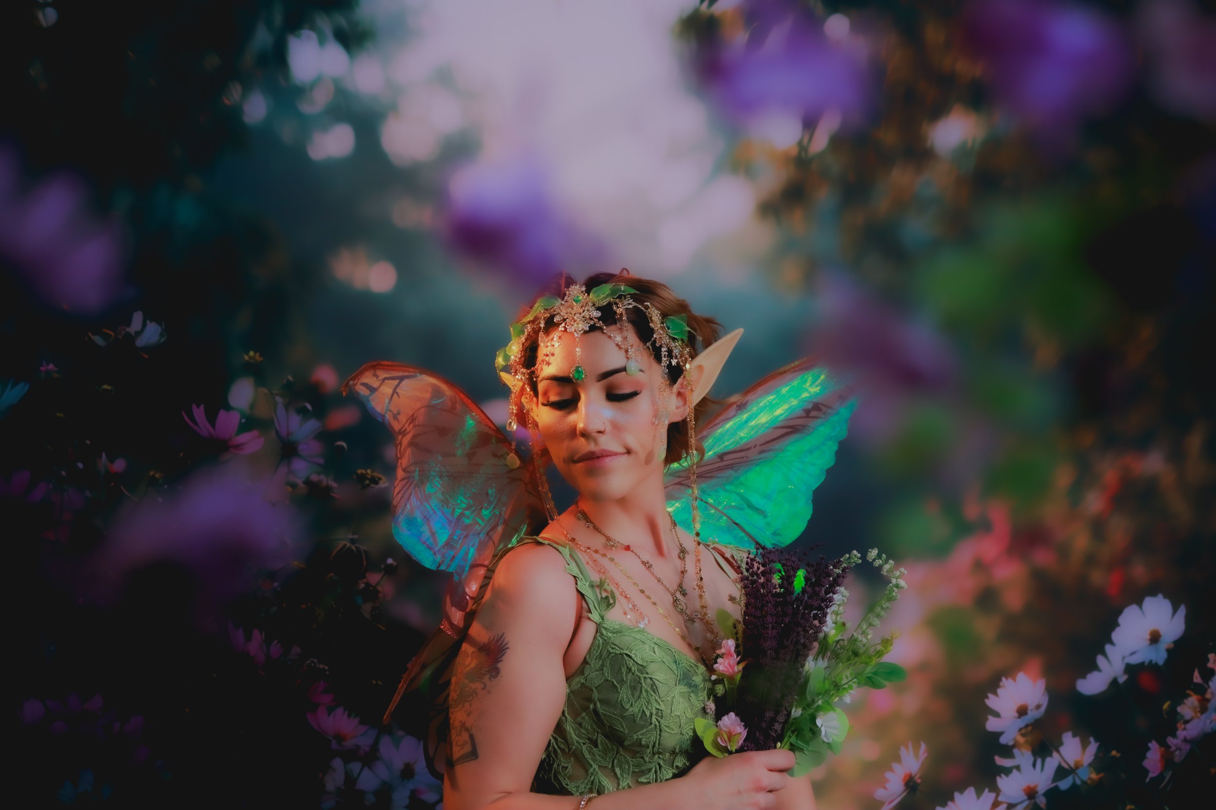 A woman dressed as a fantasy fairy with green lace dress, fairy wings, elf ears, and decorative jewelry holding a bouquet of purple flowers, standing in a garden among purple and pink flowers