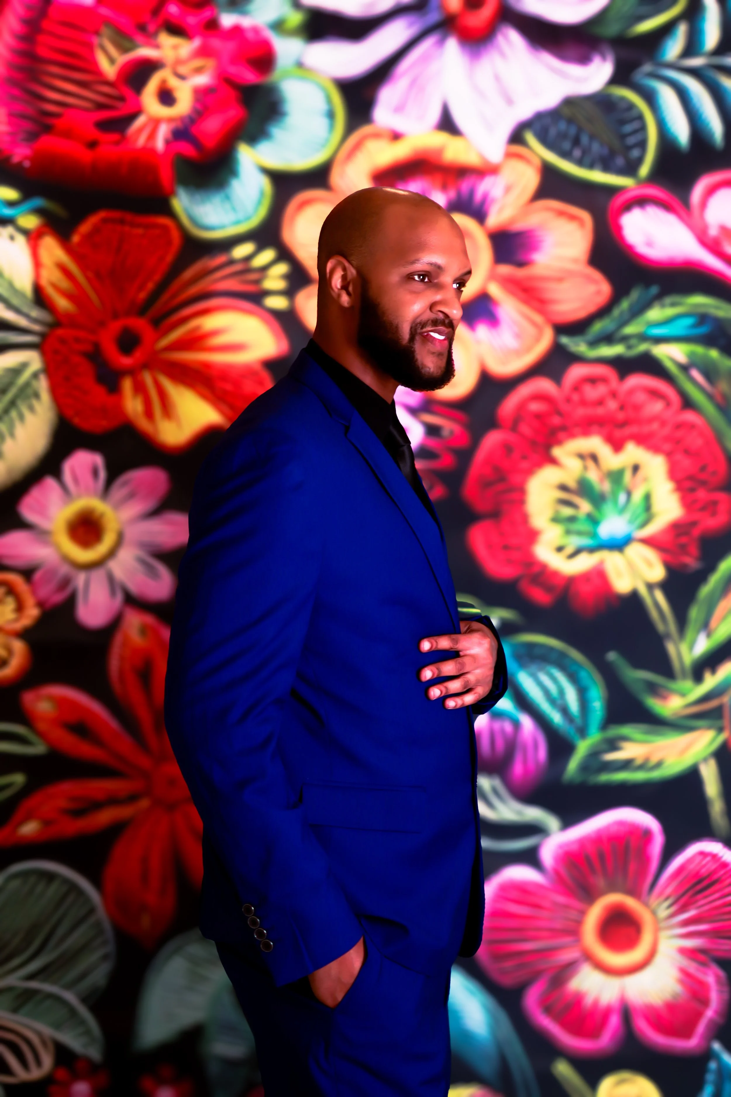 A man in a blue suit with a beard is standing in front of a colorful floral artwork background.