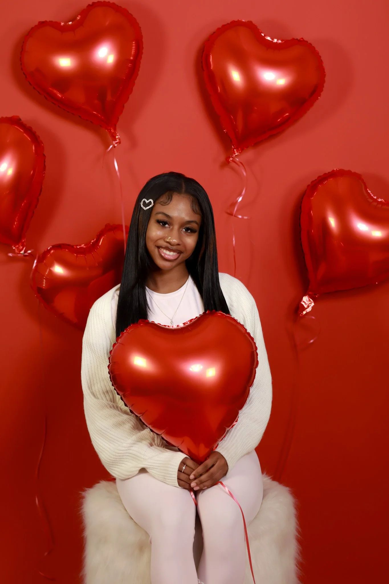 A woman sitting on a white furry stool, holding a red heart-shaped balloon, surrounded by several other red heart-shaped balloons against a red background, celebrating Valentine's Day.