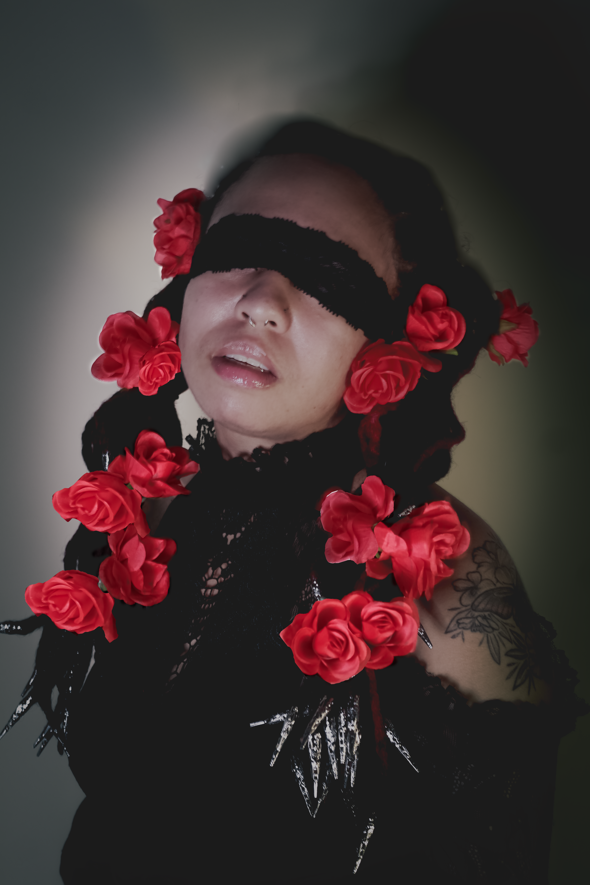 Person with a blindfold made of black lace over eyes, adorned with red roses in hair, black lace clothing, and tattoos on shoulder, in a dark, muted background.