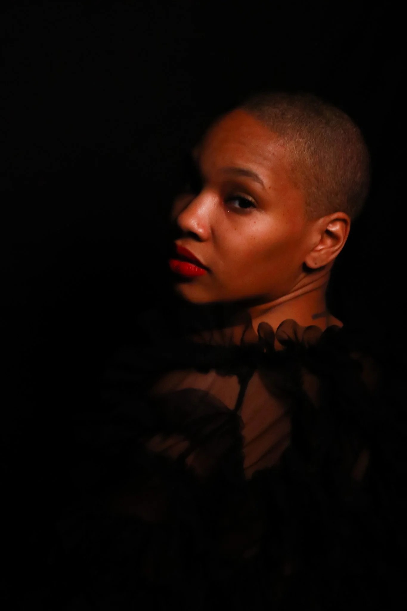 A woman with a shaved head, wearing red lipstick, in a dark setting.