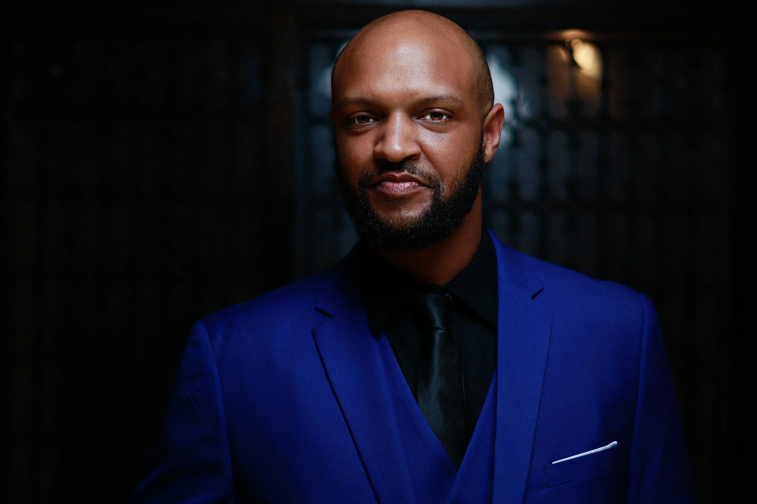 A man with a bald head and beard wearing a blue suit, black shirt, and tie, stands in a dark setting with a faint background light.