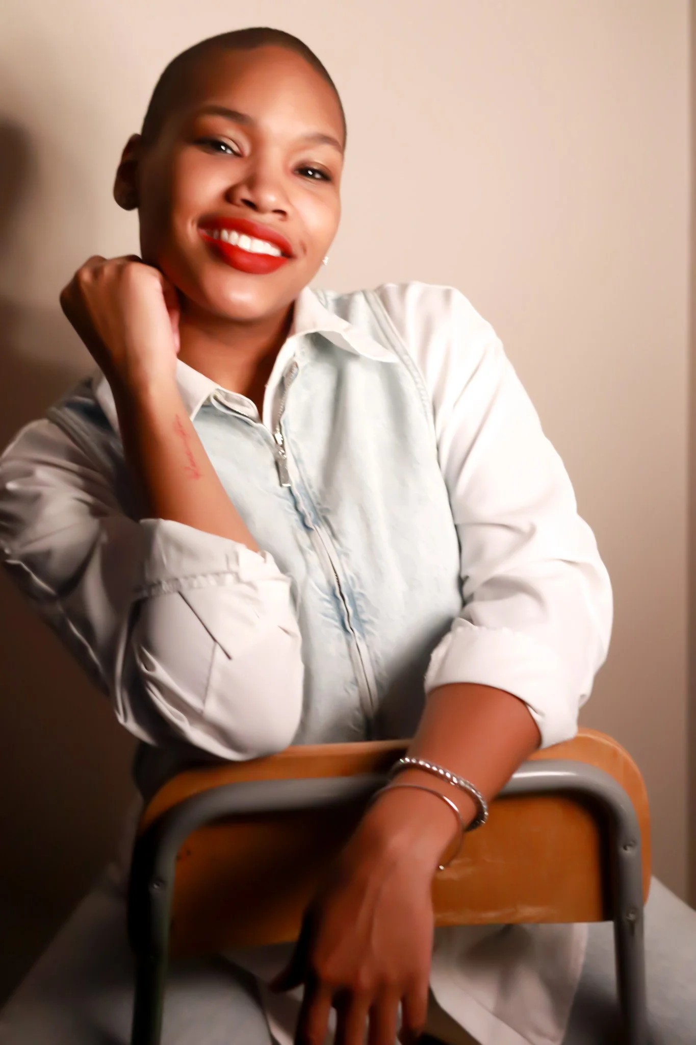 A woman with short hair, smiling, wearing a white zip-up jacket and red lipstick, sitting at a desk with her arm resting on the back of a chair.