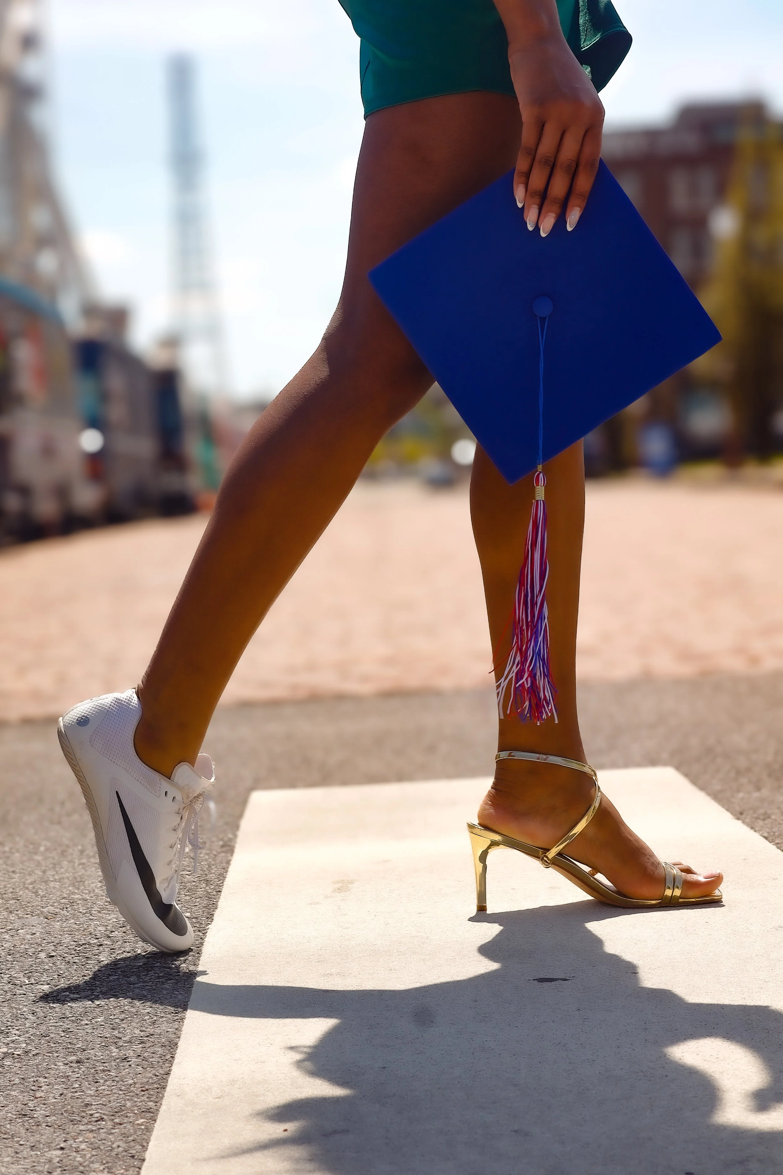 A person standing outdoors with one foot in a white sneaker and the other in a gold high-heeled sandal, holding a blue graduation cap with a multicolored tassel, indicating a graduation celebration.
