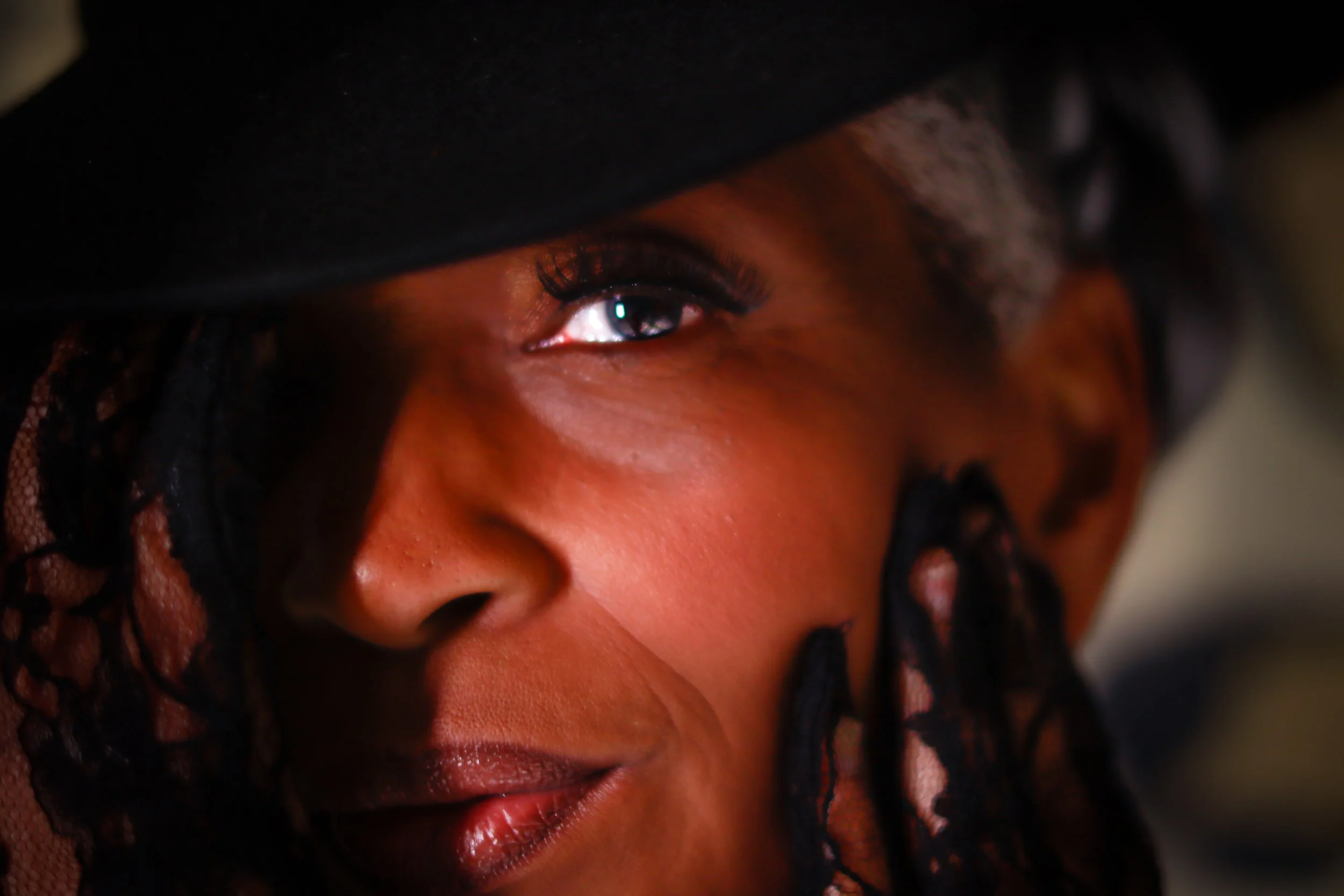 Close-up portrait of a woman with dark skin, wearing a black lace hat and makeup, with one eye partially visible.