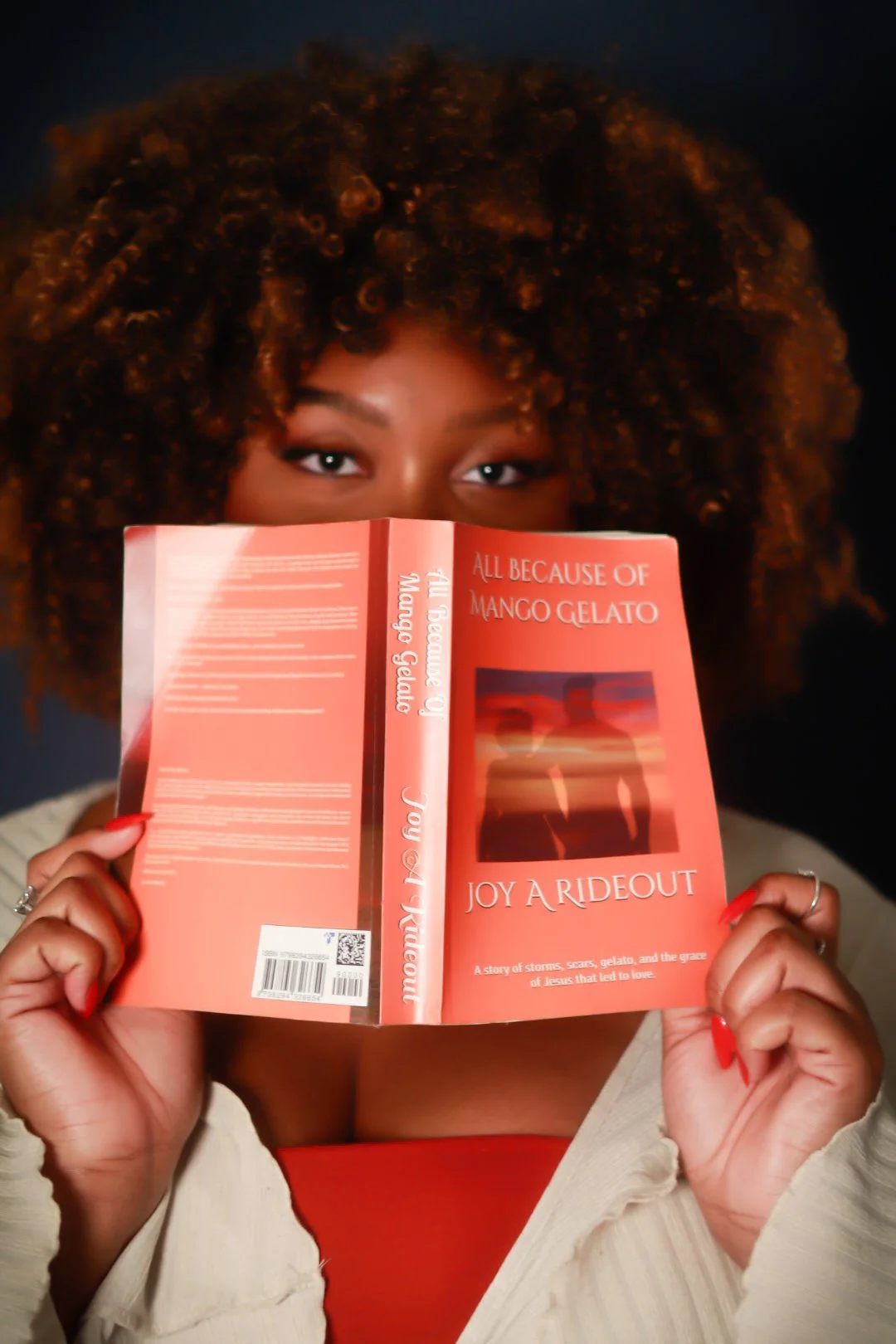 A woman with curly hair holding a red book titled 'All Because of Mango Gelato' and 'Joy a Rideout' in front of her face, revealing only her eyes, against a dark background.