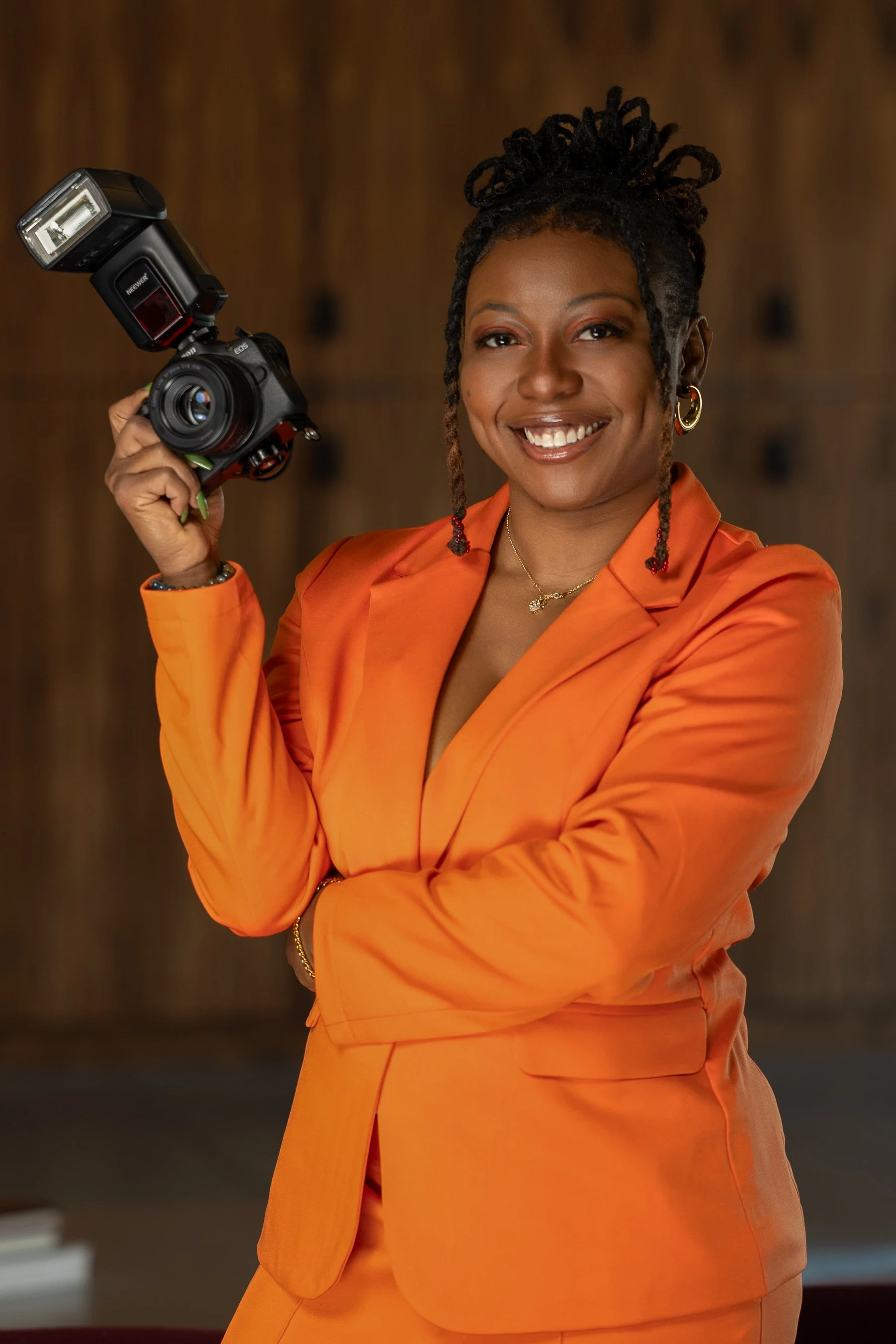 A woman with locs wearing an orange blazer and pants, smiling while holding a camera with a flash in front of a wooden background.