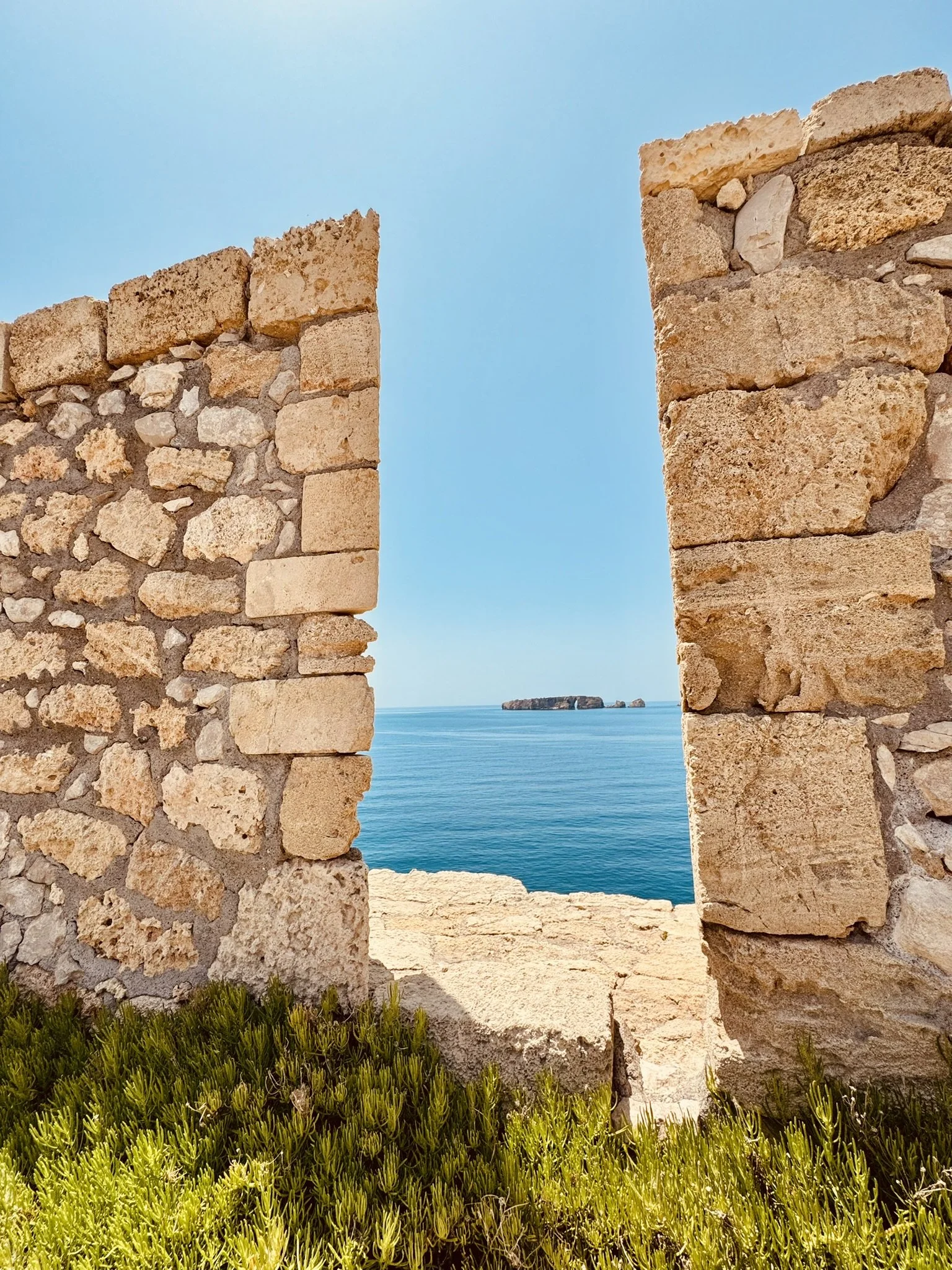 Steinmauer mit Fensteröffnung, Blick auf das Meer und eine kleine Insel in der Ferne, mit grünem Pflanzen im Vordergrund, am Tag mit klarem Himmel.
