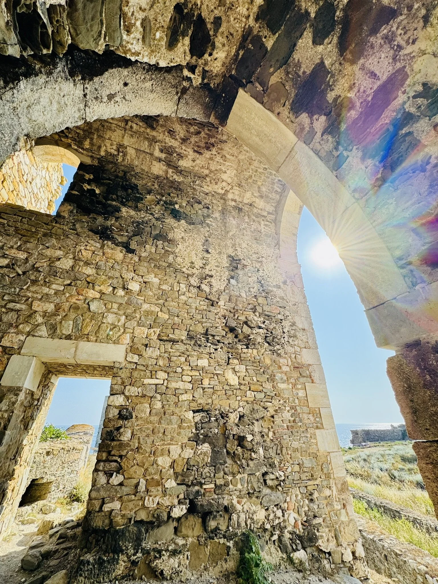 Alte Steinmauer mit Bögen und Fensteröffnungen unter strahlendem Sonnenlicht, Blick auf das blaue Himmel und das Meer im Hintergrund.