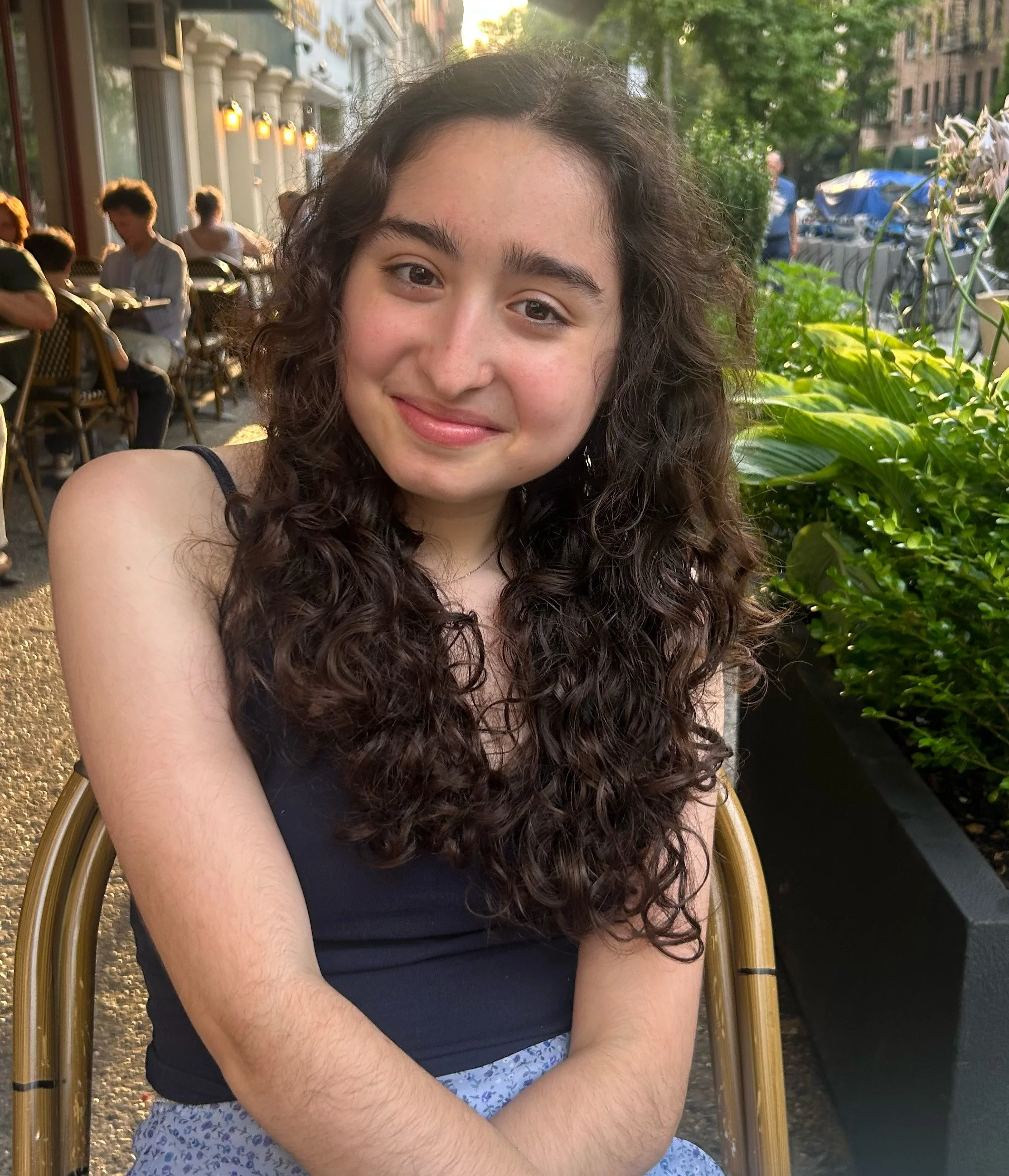 A young woman with curly brown hair smiling on an outdoor cafe patio.