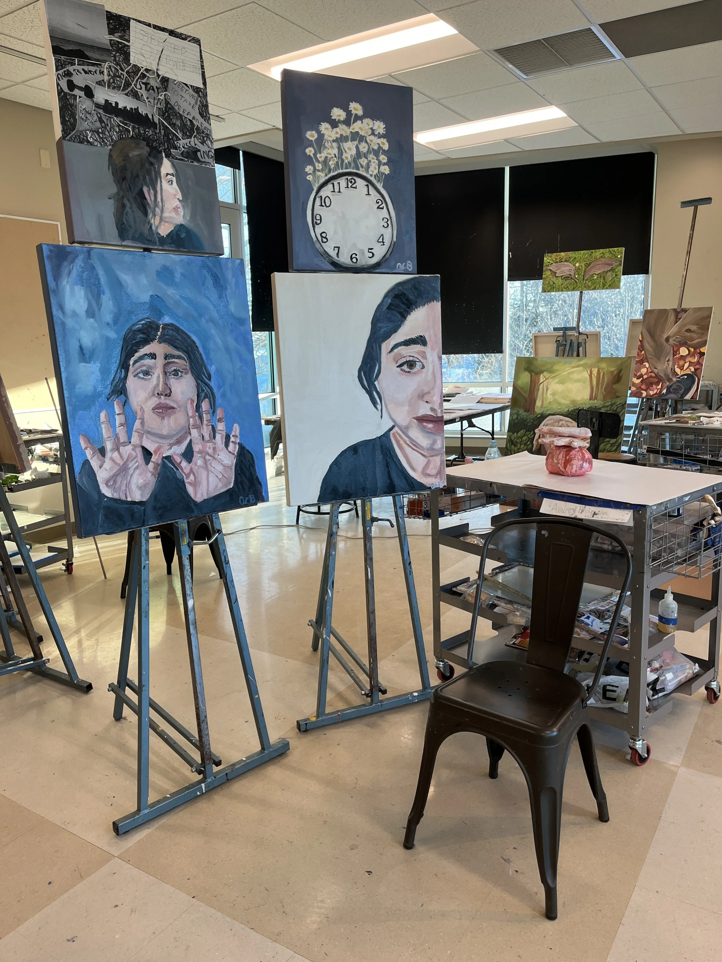 A room with several easel paintings of women, a clock with daisy flowers, and nature scenes. Art supplies and a black chair are also visible.