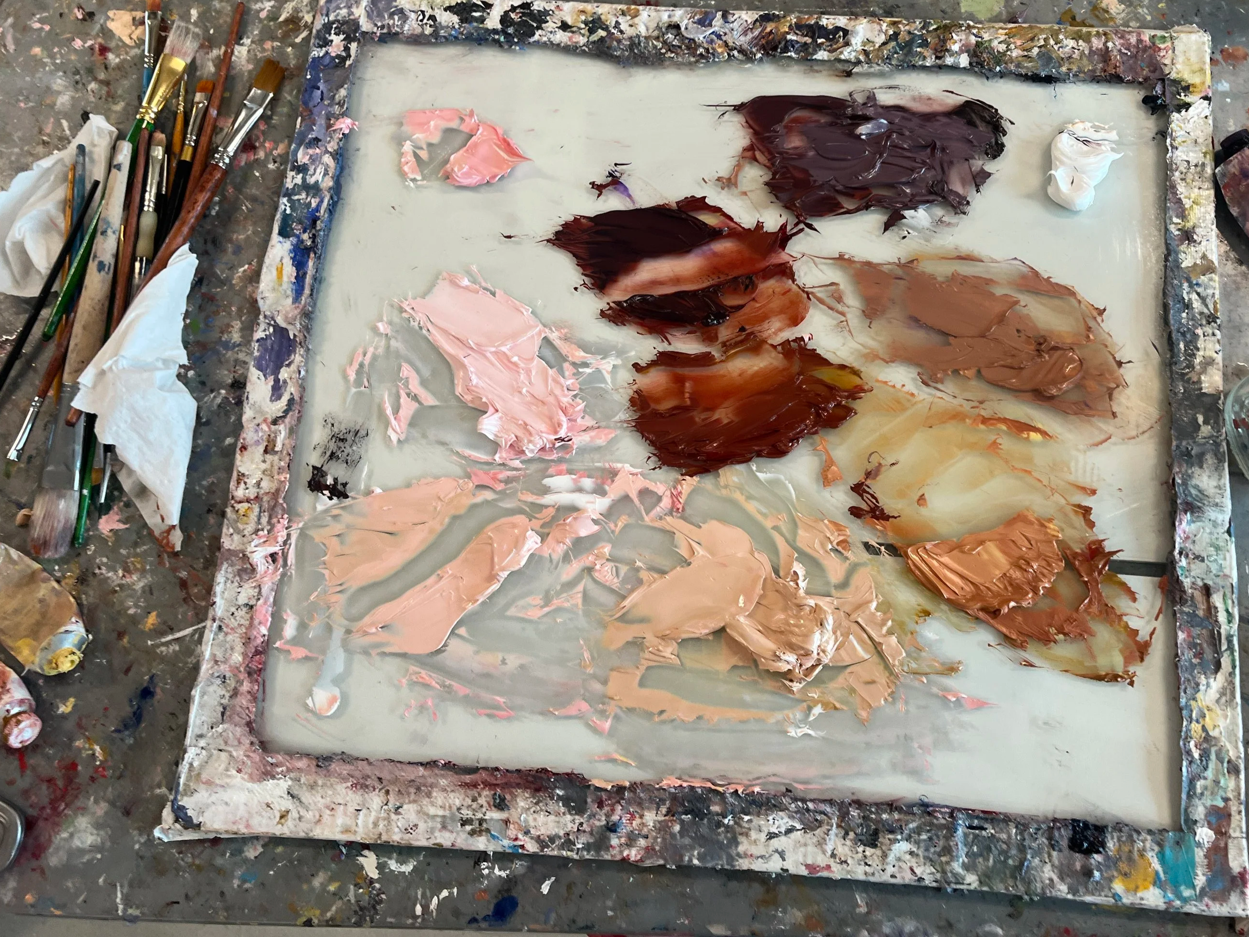 Artist's palette with various shades of pink, brown, and beige oil paints, surrounded by paintbrushes and painting supplies on a messy art table.