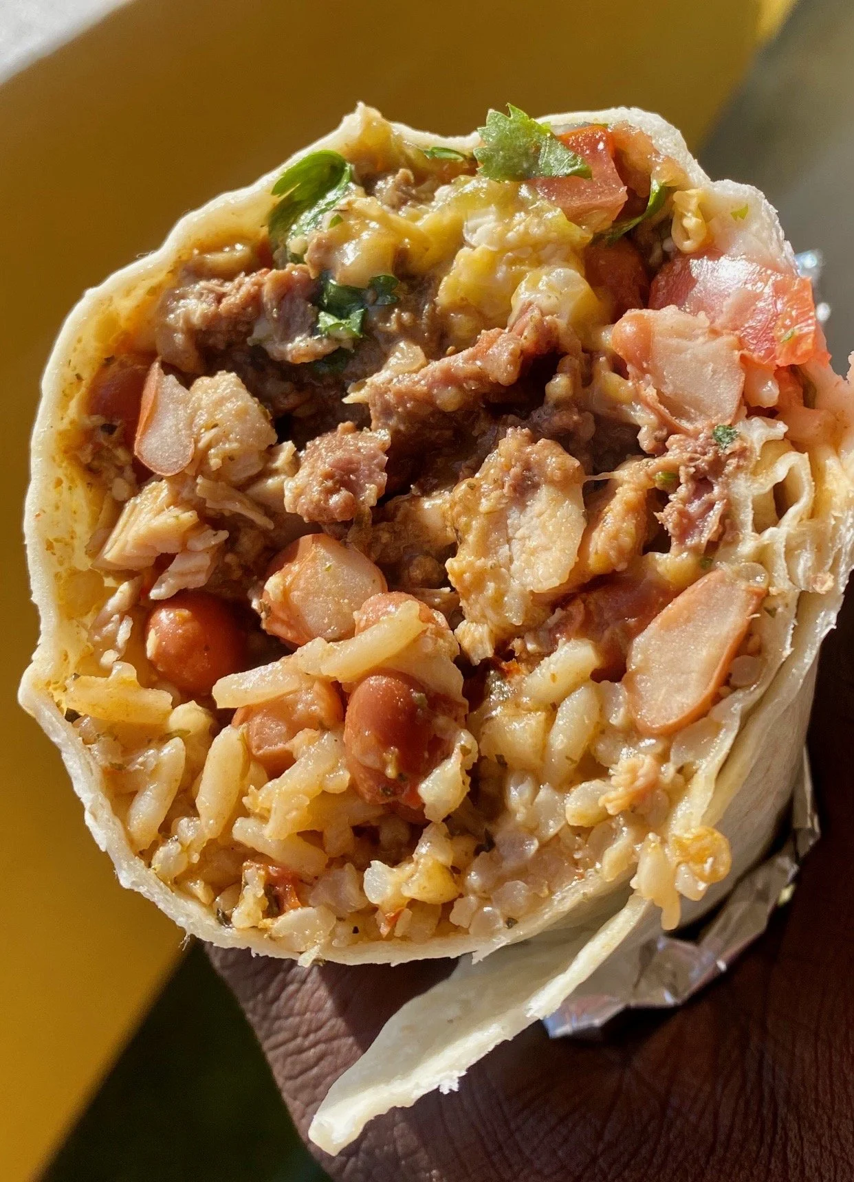 Close-up of a burrito filled with rice, beans, meat, tomatoes, and cilantro, held by a person.