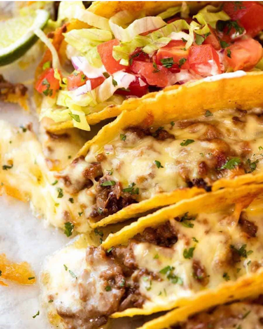 Three crispy taco shells filled with seasoned ground beef and melted cheese, served with shredded lettuce, diced tomatoes, and chopped onions topped with fresh cilantro, alongside lime wedges and salsa.