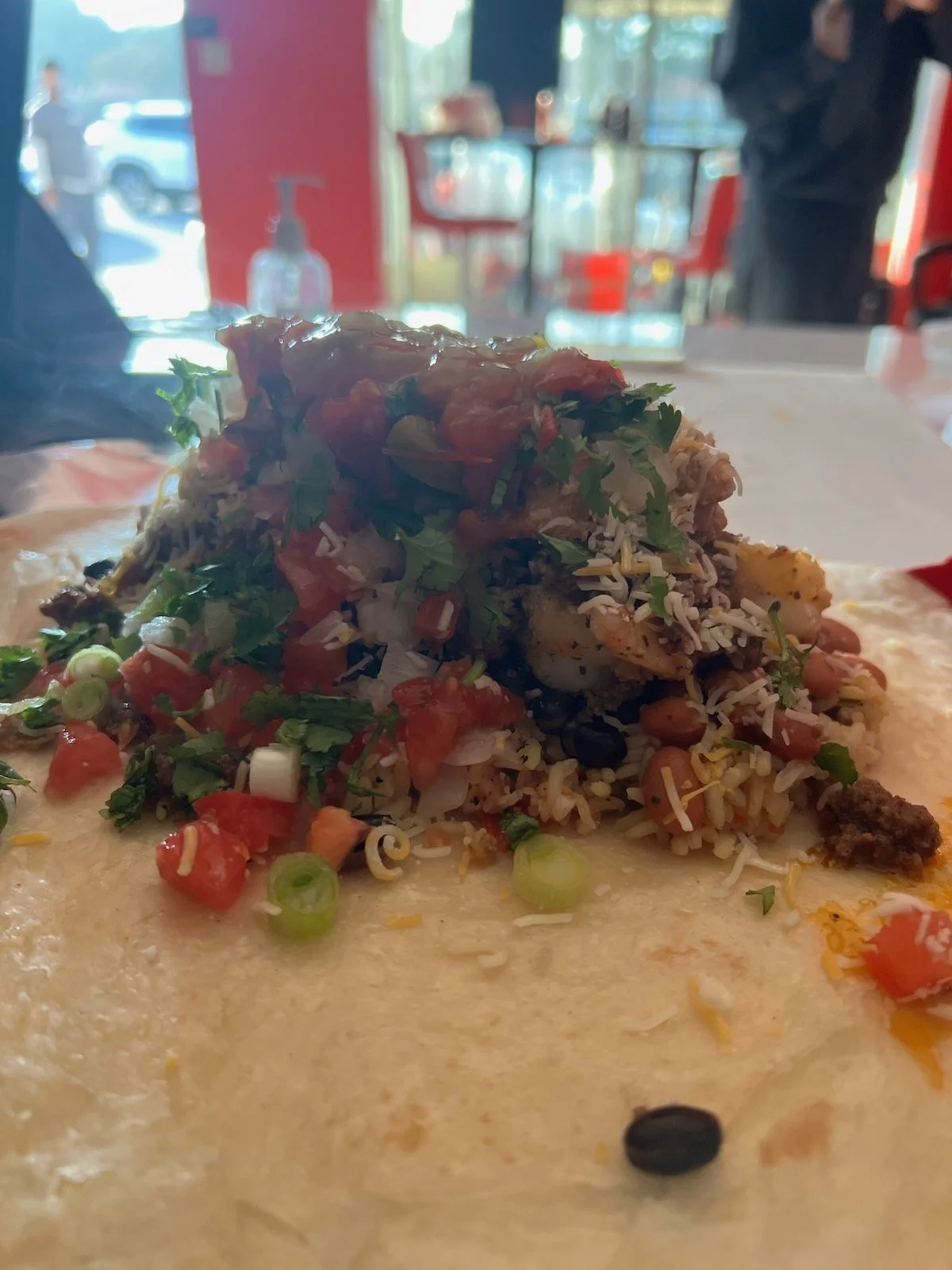 Close-up of a loaded taco with rice, beans, salsa, shredded cheese, and cilantro on a paper plate inside a restaurant with blurred background.