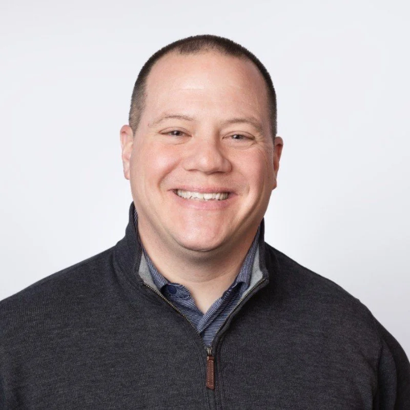 Smiling man with short hair wearing a dark zip-up sweater over a collared shirt, against a plain white background.