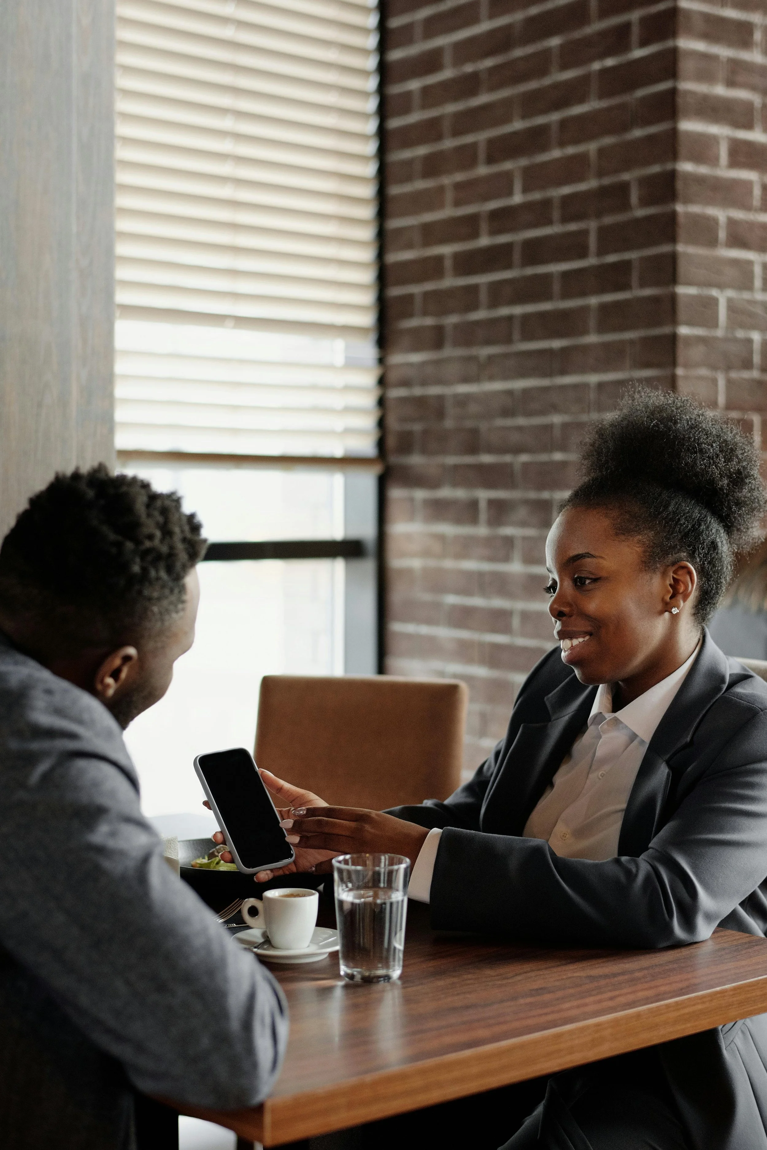 Two people, a woman and a man, sitting at a wooden table in a cafe, with the woman showing something on her phone to the man, who is smiling.