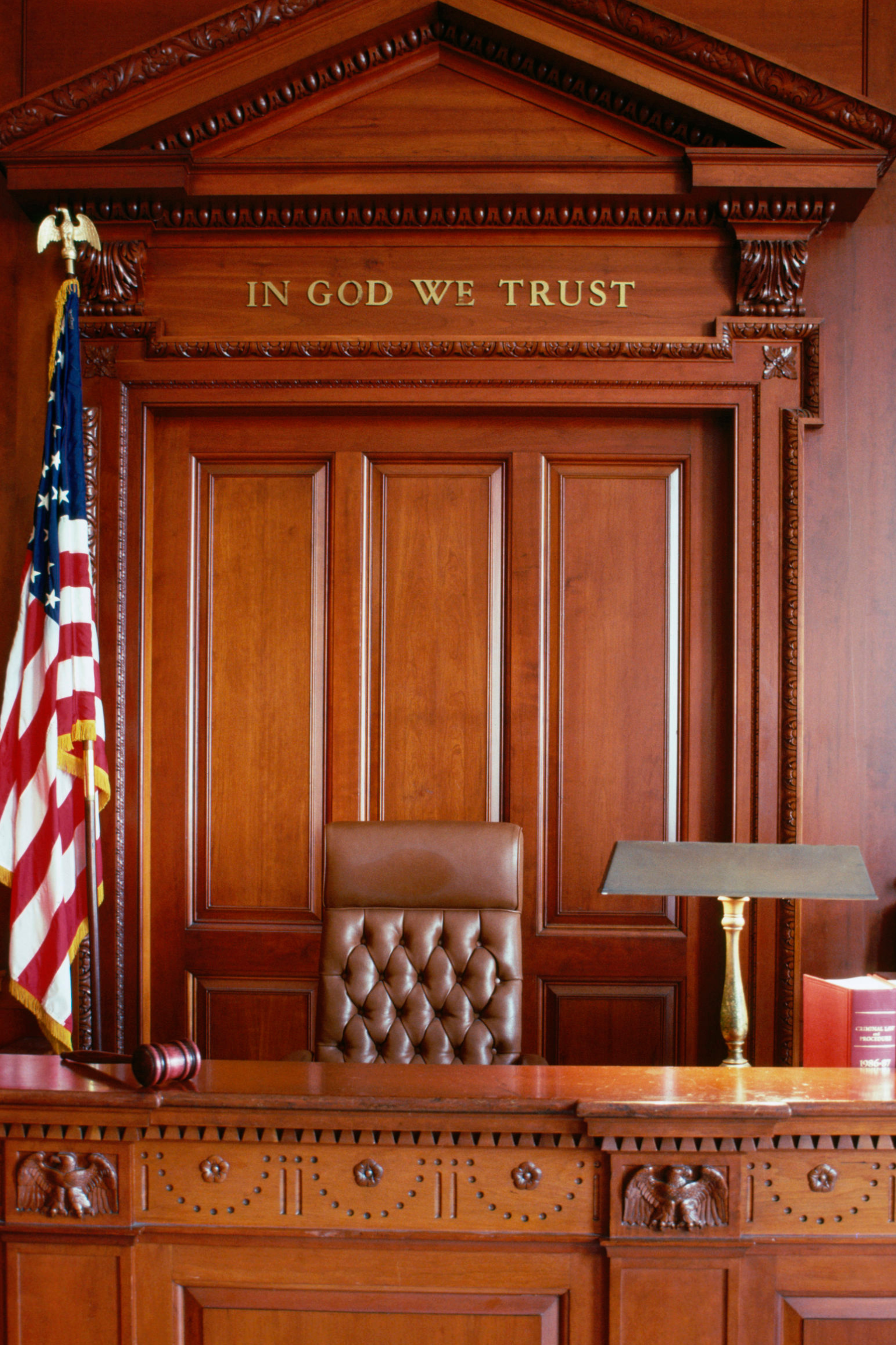 A judge's bench in a courtroom with a wooden gavel, a table lamp, a legal book, and an American flag on the side. The backdrop features a large wooden panel with the words "IN GOD WE TRUST" engraved above it.
