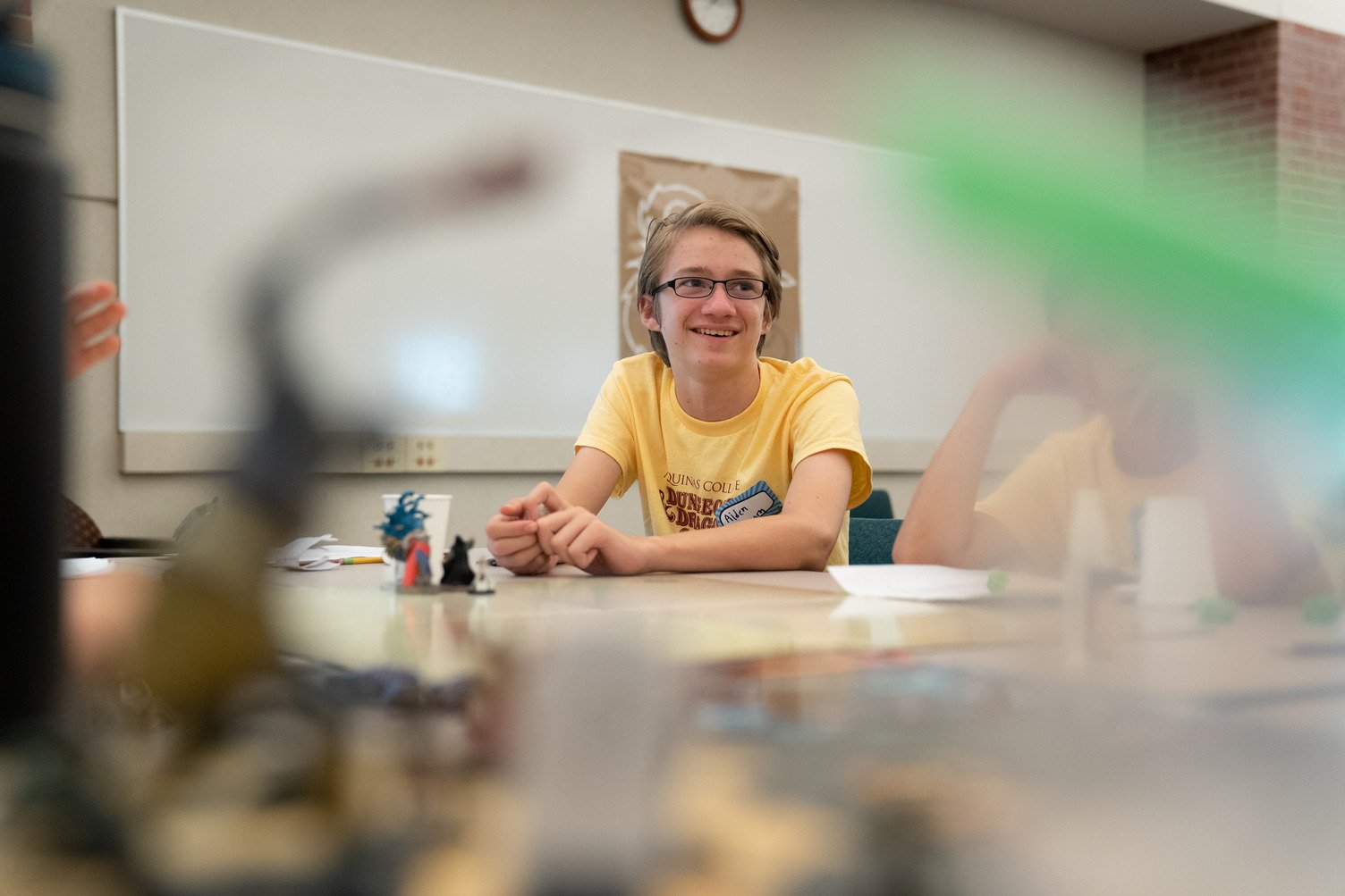 A young man with glasses and a yellow T-shirt sitting at a table in a classroom, smiling with his hands clasped, surrounded by blurred objects on the table and another person partially visible to the right.