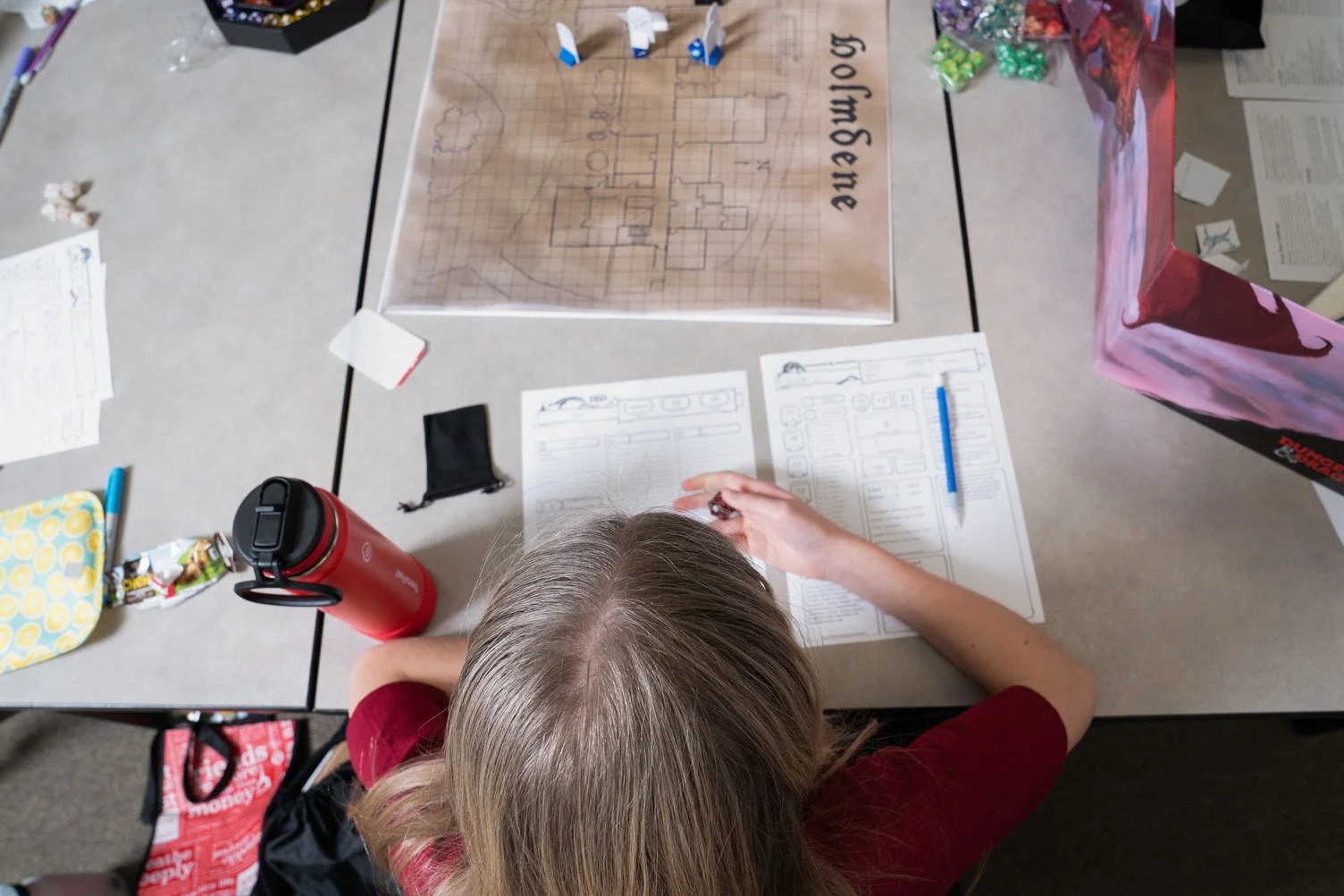 A person with blonde hair and a red shirt is sitting at a table, writing on a sheet of paper with diagrams and text. The table has various items including a red water bottle, a yellow patterned pouch, a black pouch, a blue pen, and a large gaming map