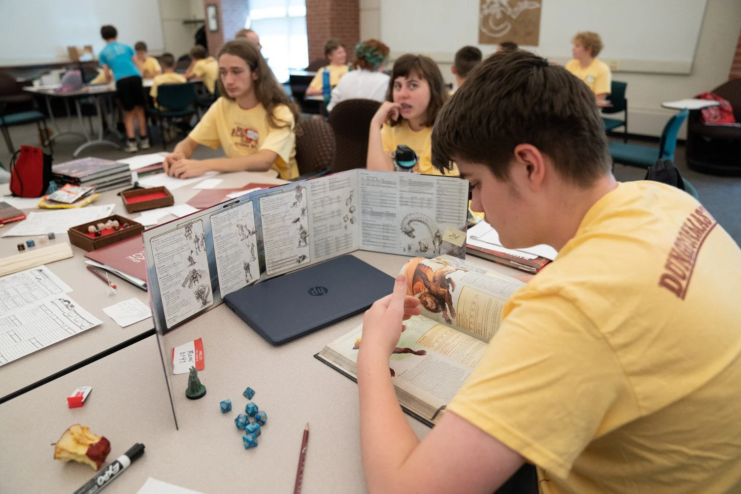 Group of students in yellow shirts sitting around a table with gaming and reference books, dice, and writing materials, likely in a classroom or club setting.