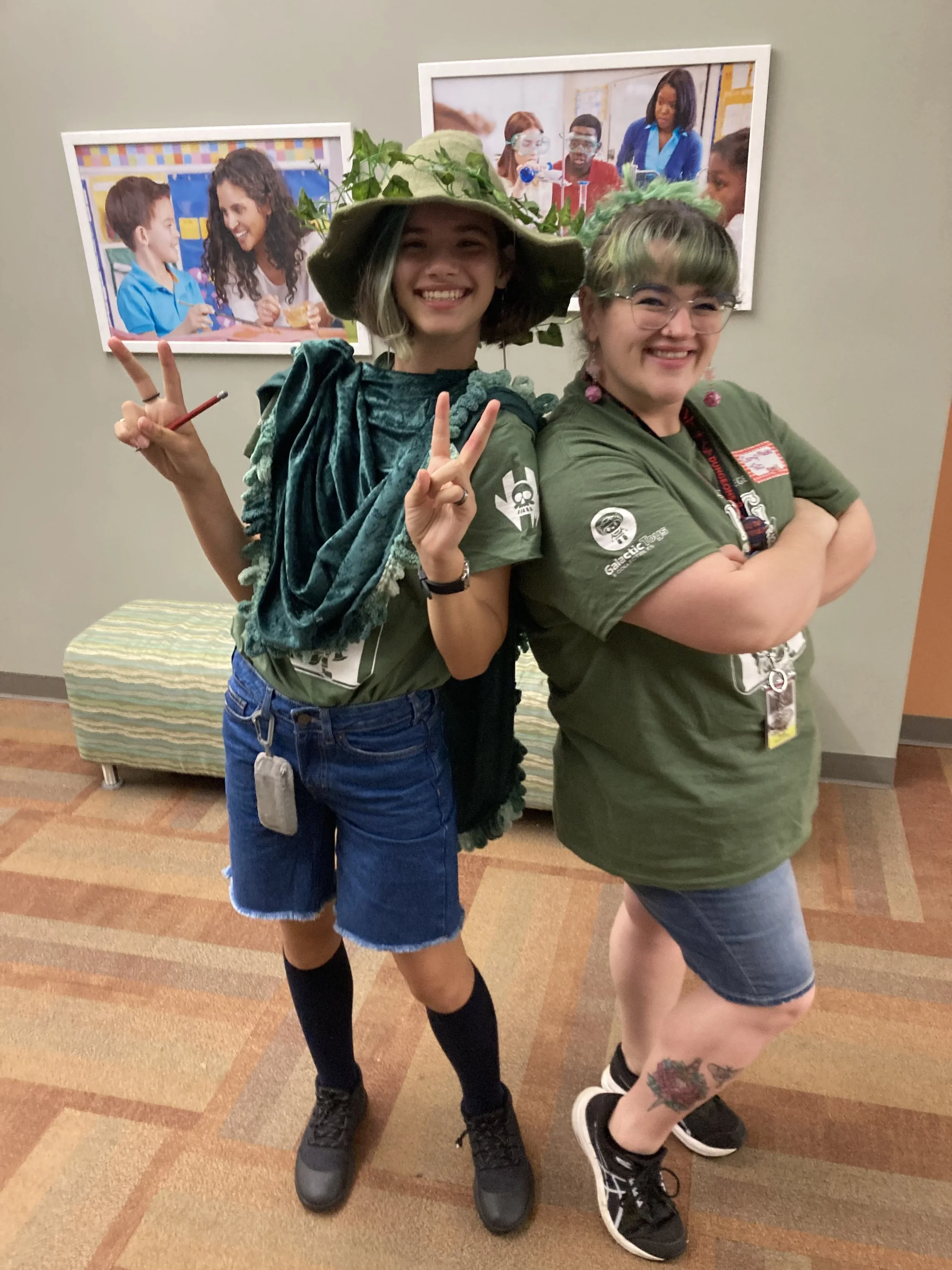 Two women in green shirts posing back-to-back with arms crossed, smiling and making peace signs, standing indoors in front of framed pictures of children and teachers.