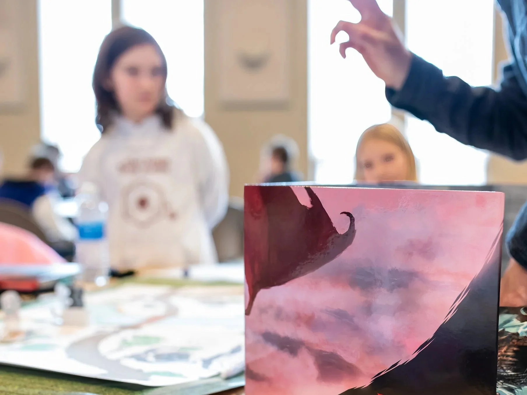 A tabletop game session with a colorful card showing a mountainous landscape and a silhouette of a dragon in a pink sky, with blurred children and an adult in the background, inside a bright room.