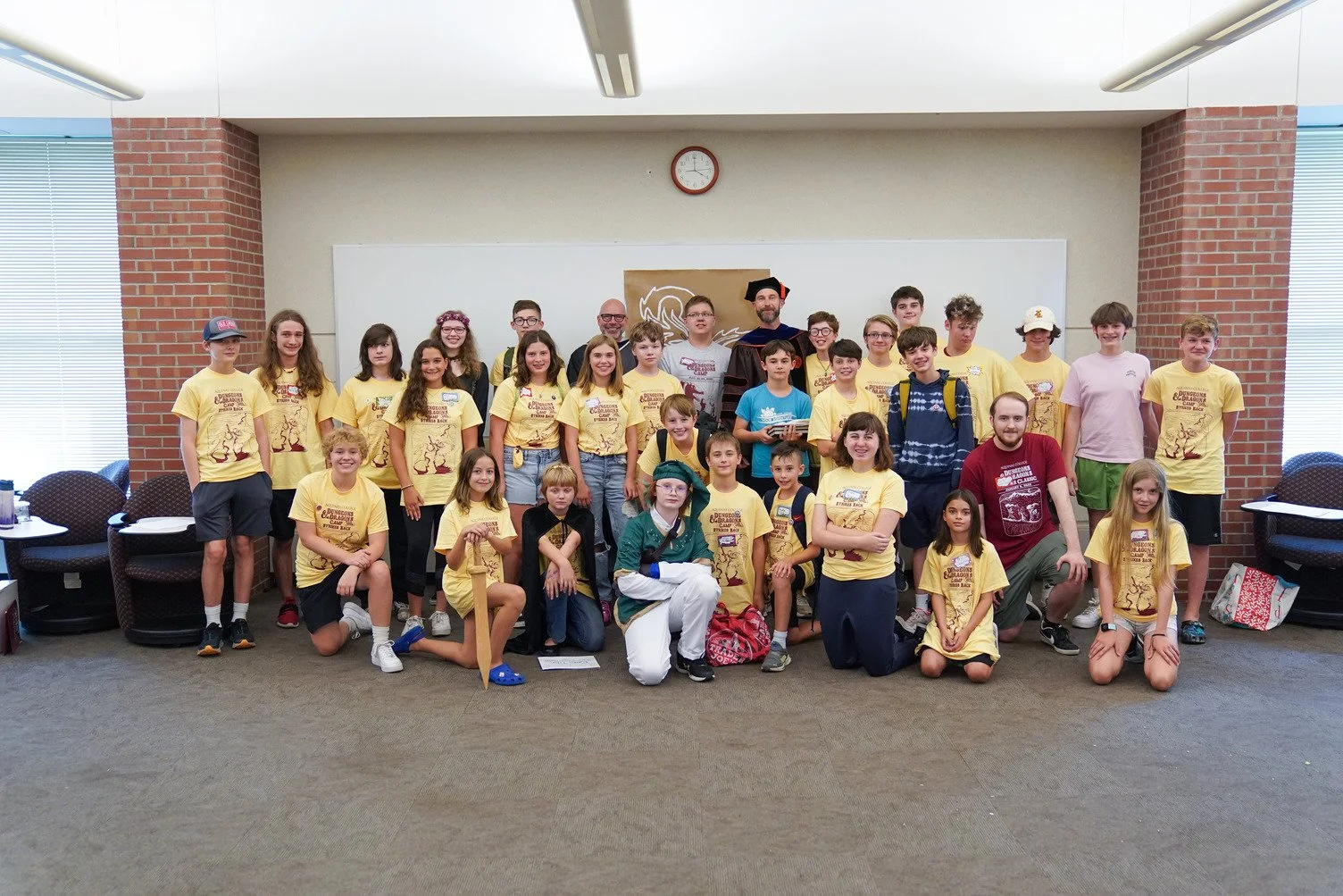 Group of children and adults on a stage at a scout event, some wearing yellow shirts with a logo, others in costumes, with a whiteboard and clock in the background.