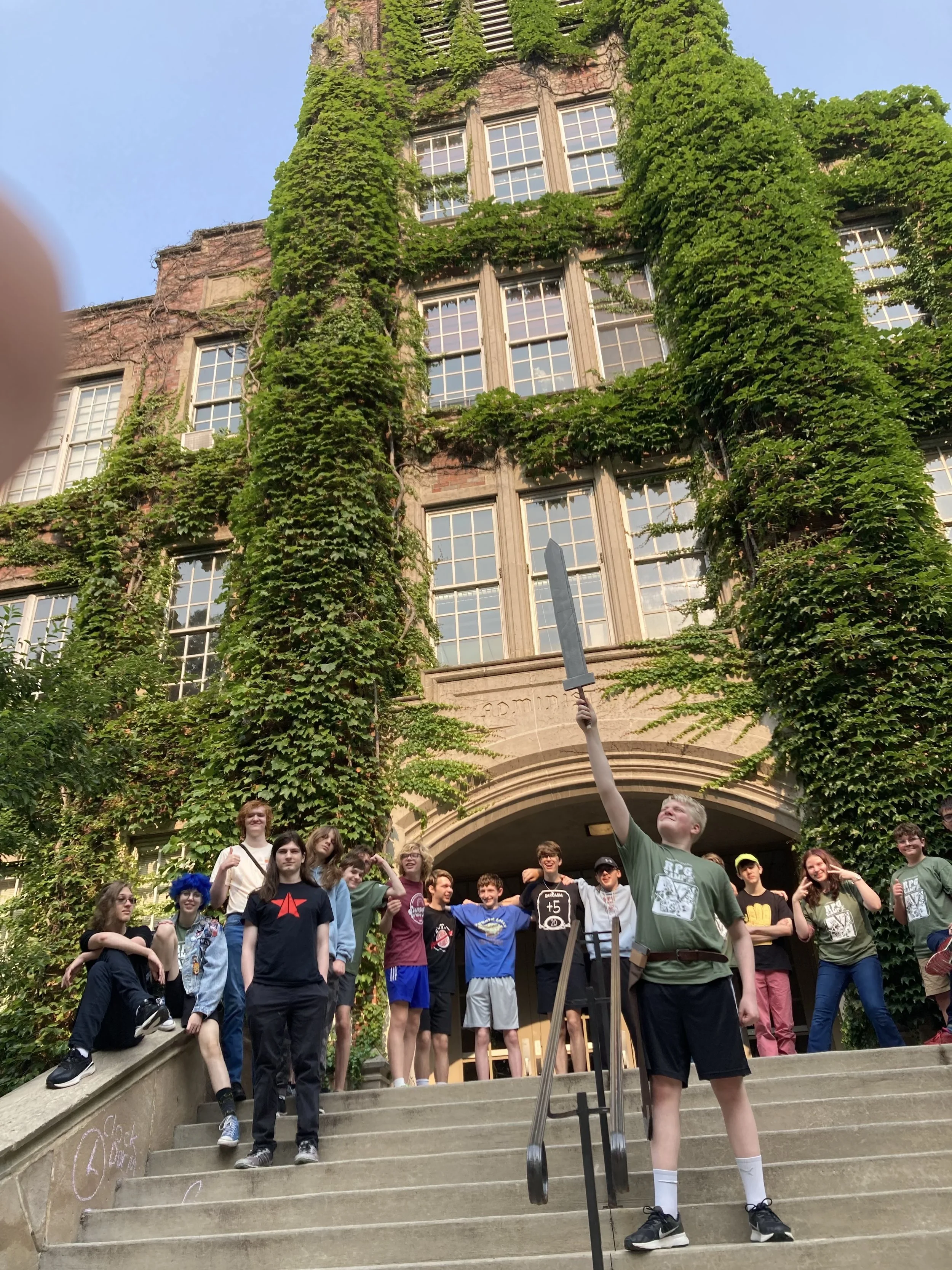 A group of kids standing on stairs outside a large building covered in green ivy. One boy in front is holding a sword upright in the air, mimicking a heroic pose. The building has tall windows and an arched entrance.